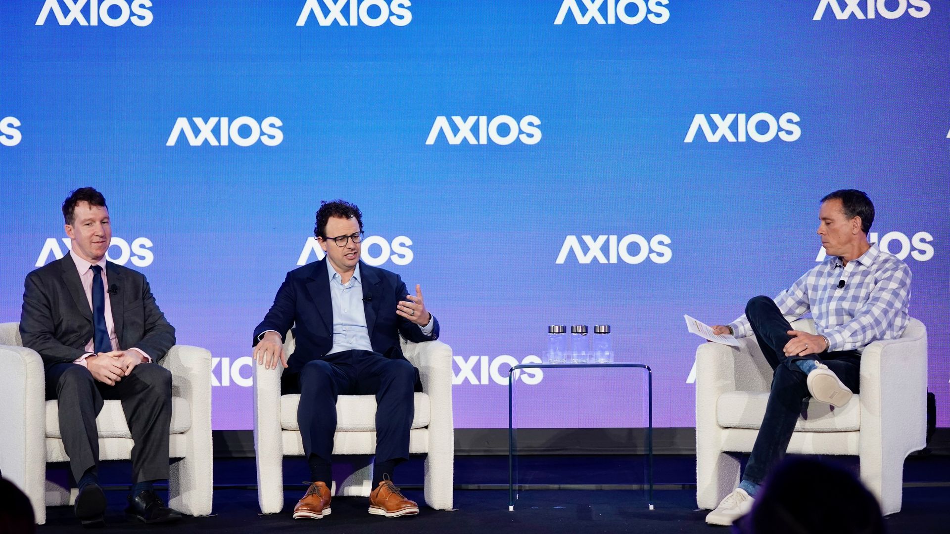 Three men seated on a stage with a blue backdrop featuring the white Axios logo, engaged in a panel discussion. One man on the right holds a paper, others are listening or speaking.