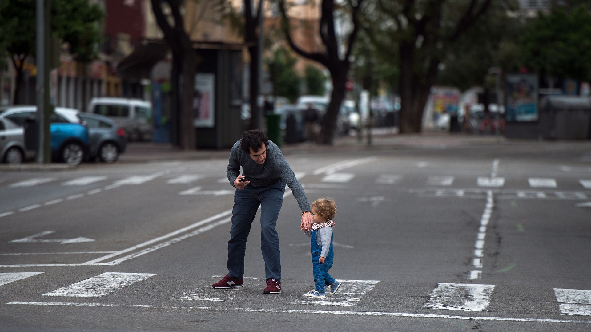 In this image, a man helps a toddler cross the street on a crosswalk in Spain
