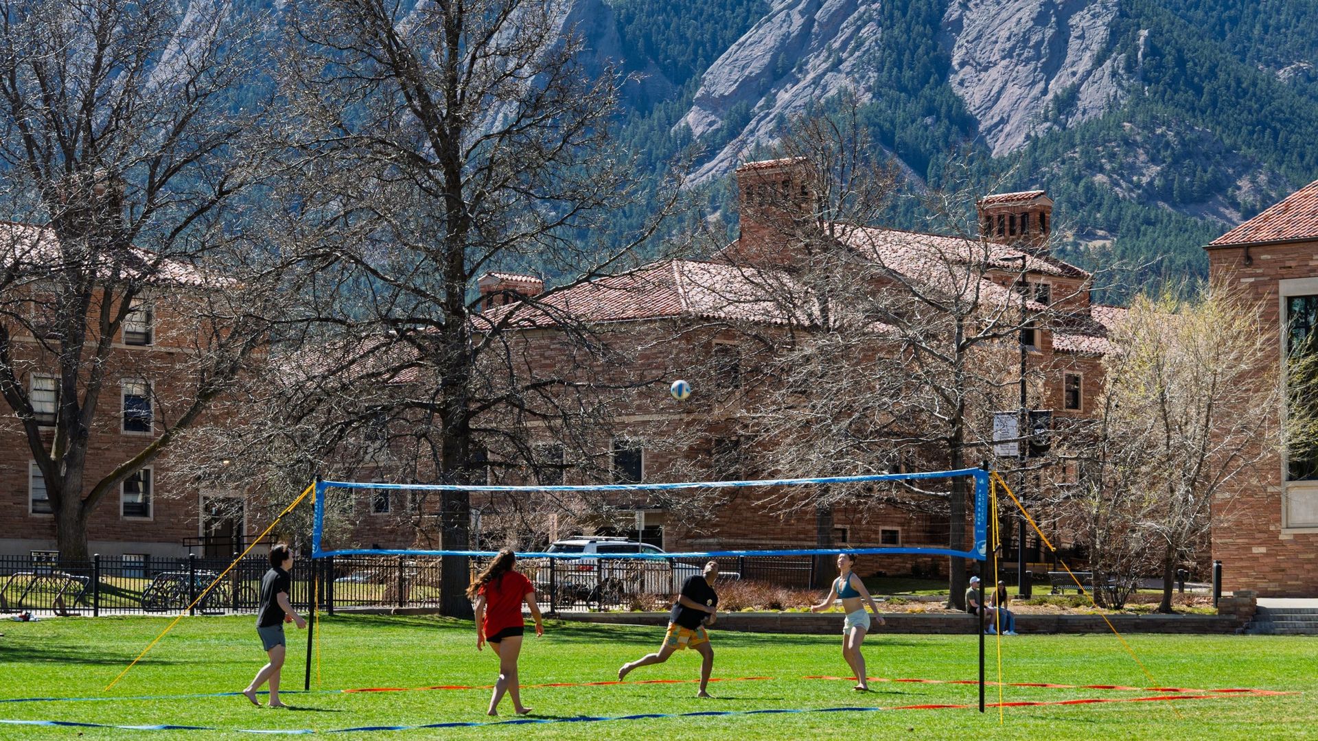 Four people play volleyball on a sunny grassy campus quad with a blue net; leafless trees, brick buildings, and rugged mountains rising behind them.