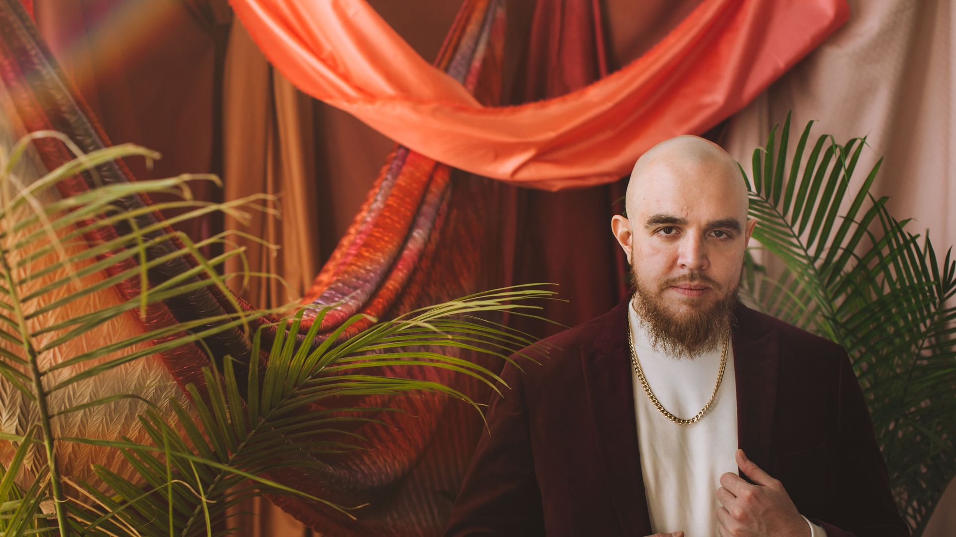 Poet José Olivarez poses for the camera with green plants behind him and red draping also behind him