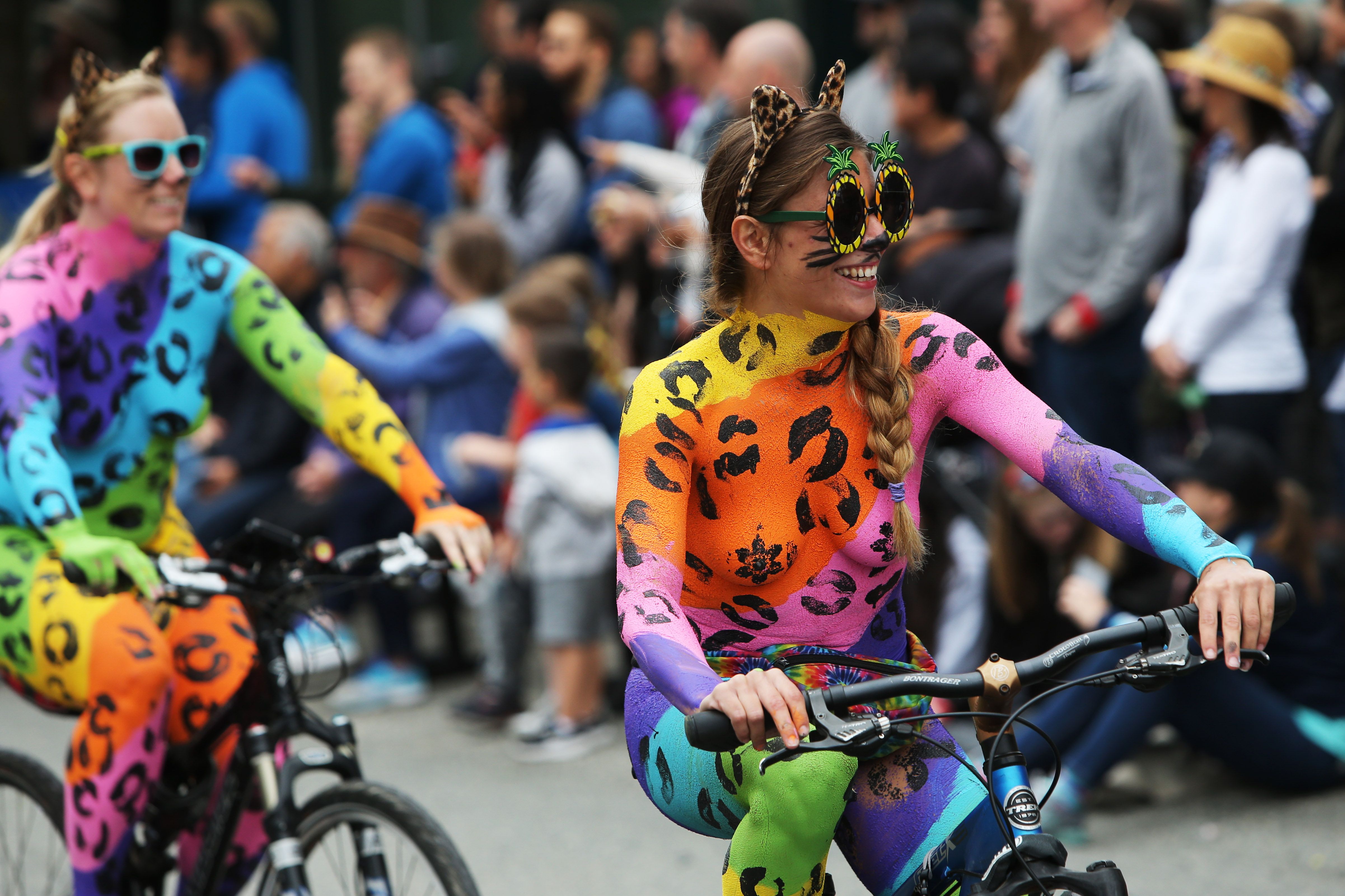 Two nude bicyclists with their skin covered by rainbow-colored body paint ride in a parade.