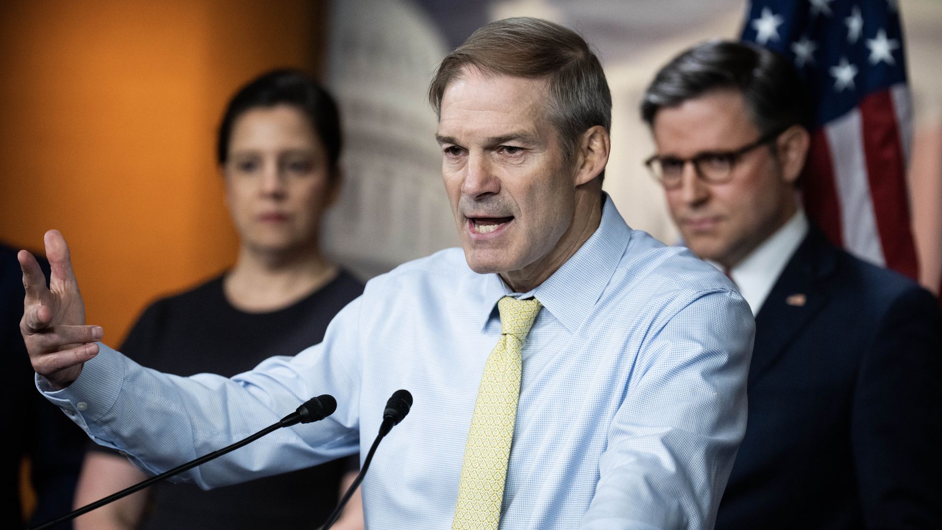 Rep. Jim Jordan, wearing a blue shirt and lime green tie, speaking at a podium while flanked by colleagues in an orange room with an American flag and a mural of the Capitol.