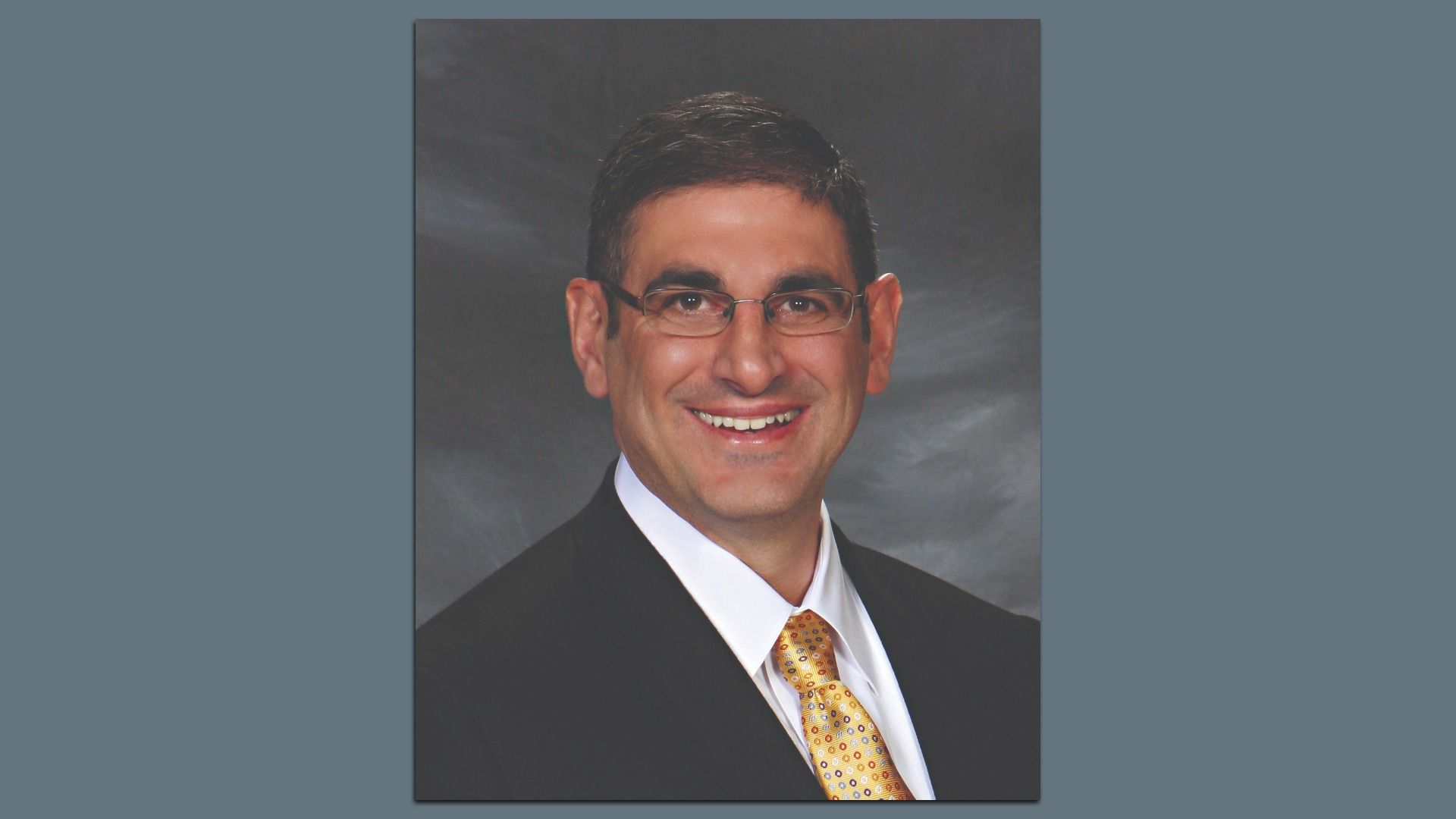 Portrait of a smiling man with short dark hair, wearing glasses, a black suit, white shirt, and yellow patterned tie, against a gray background.
