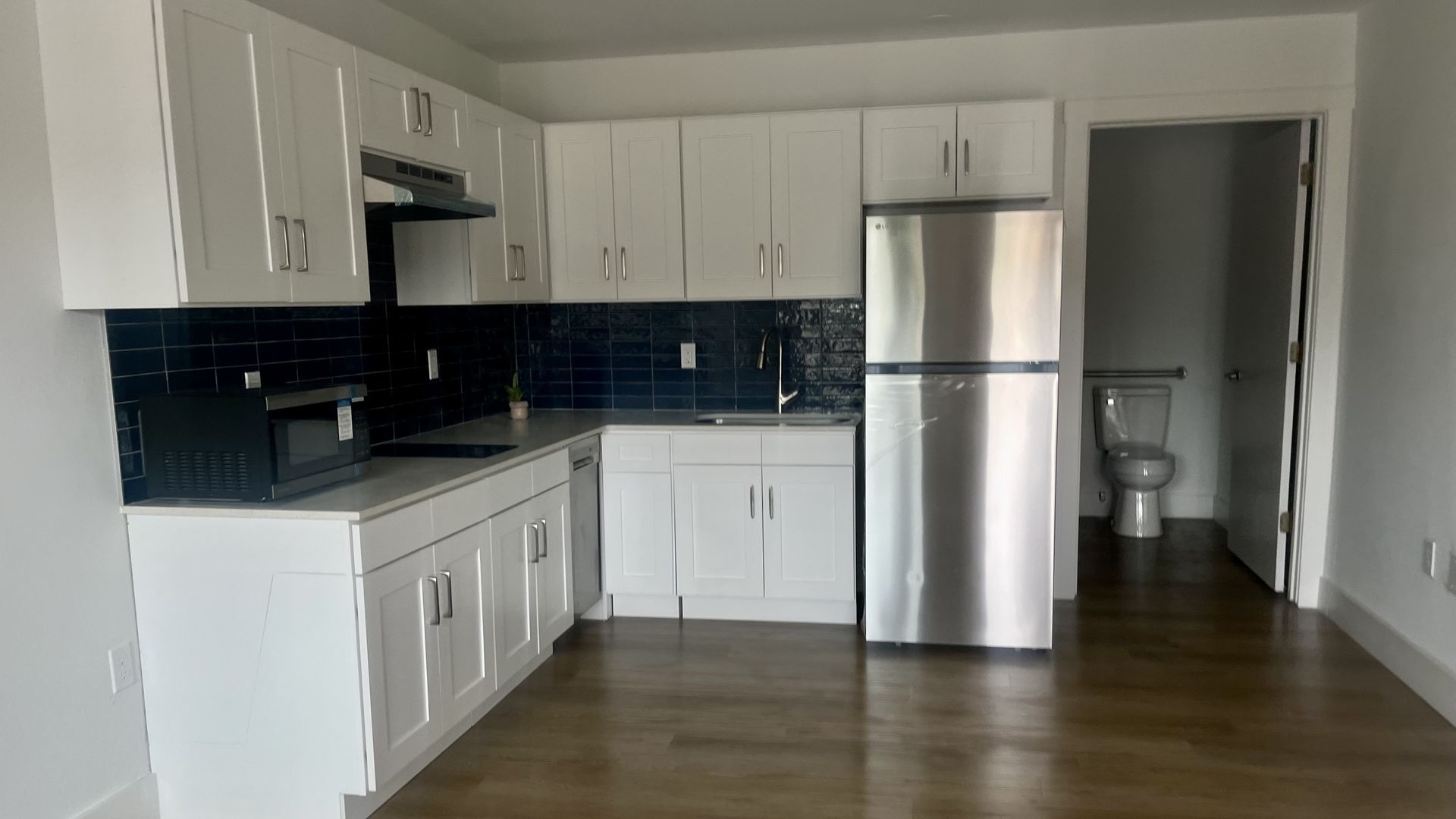 Modern kitchen with white cabinets, dark blue tiled backsplash, stainless steel refrigerator, microwave, sink, and wooden floor. Open door reveals a bathroom with toilet.
