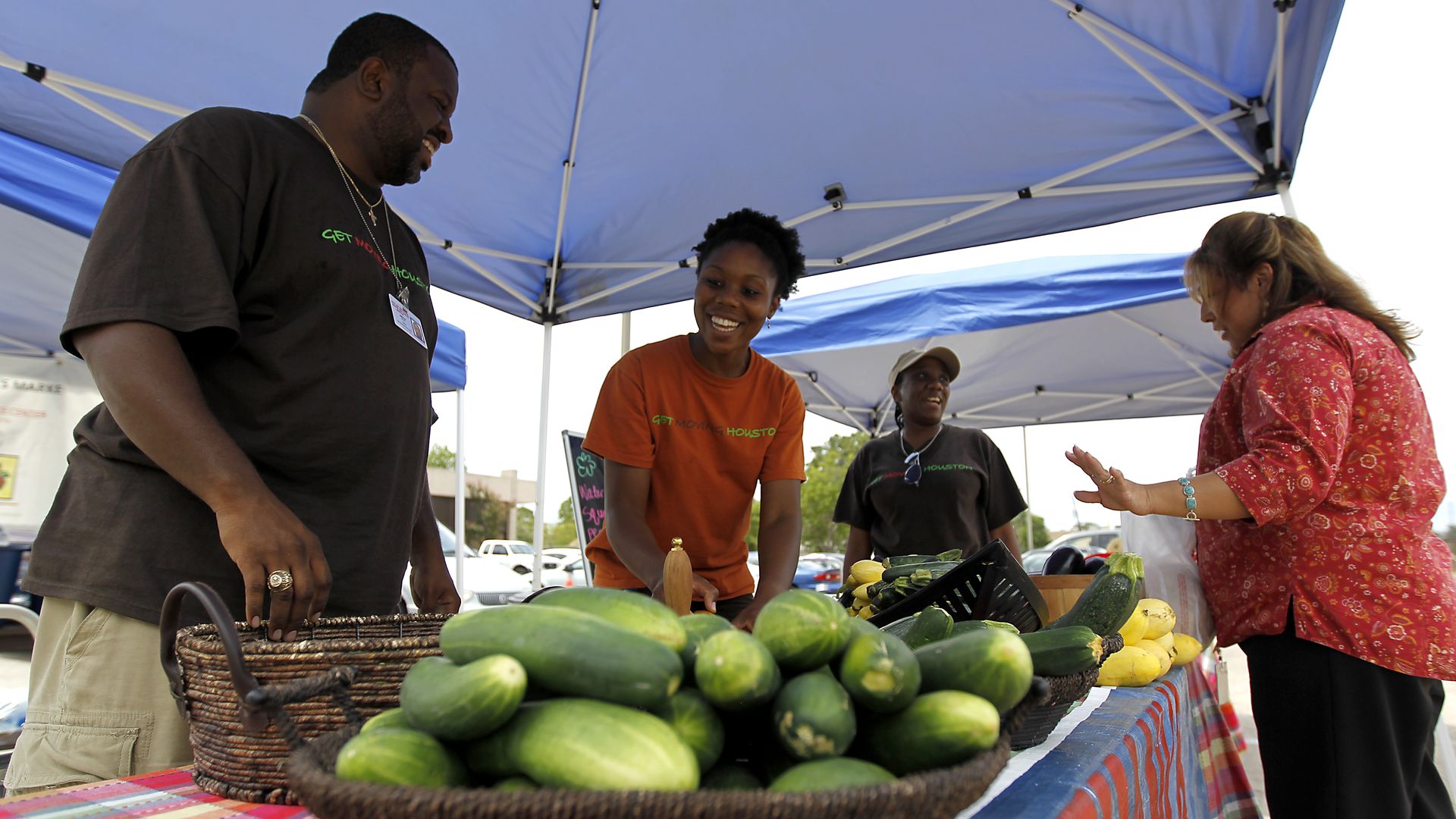 Photo of a produce stand with cucumbers, and people talking to the vendors. 