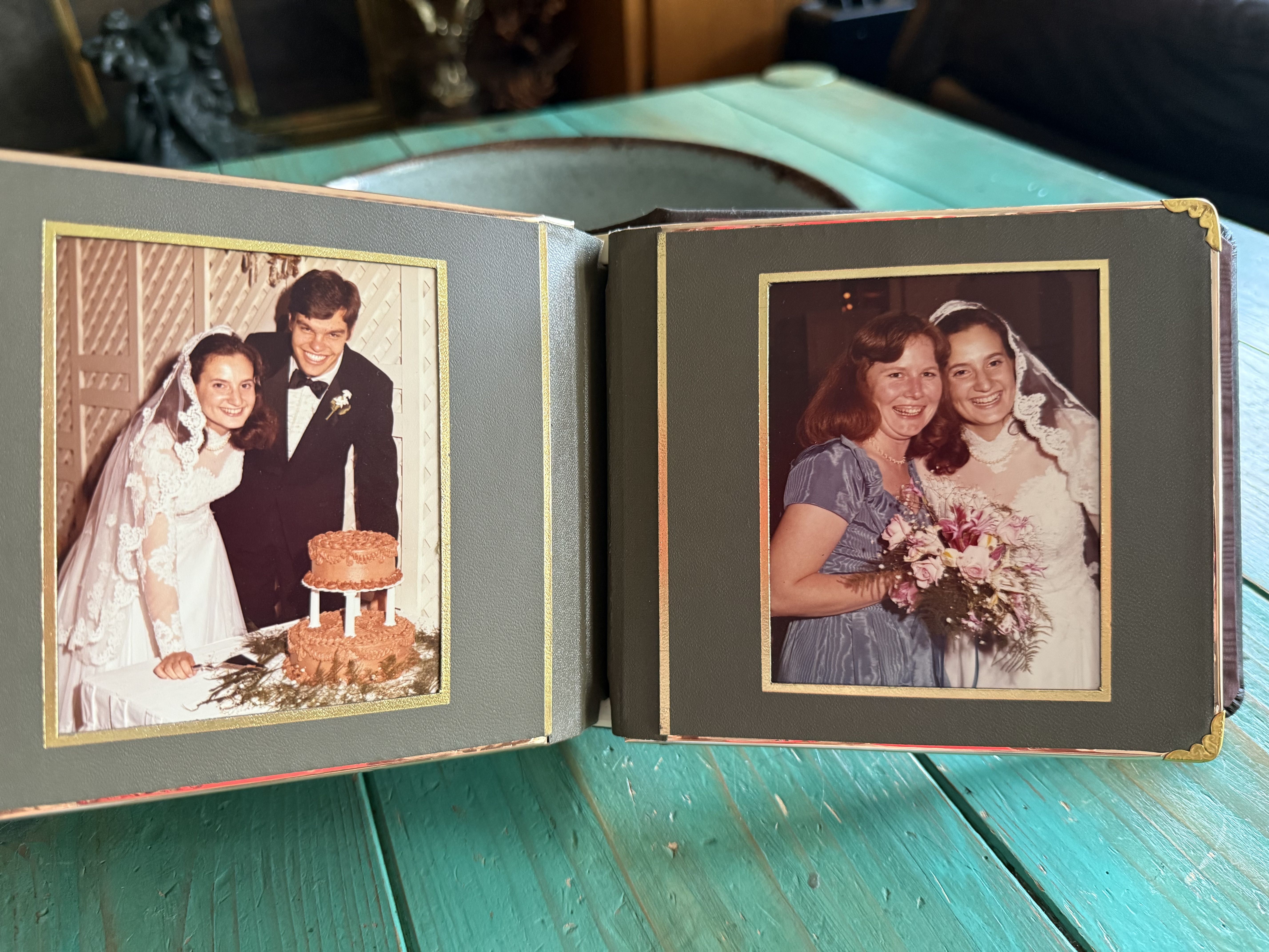 Open photo album on a teal wooden table showing two wedding photos: left of bride and groom cutting chocolate cake, right of bride with woman in blue dress holding bouquet.