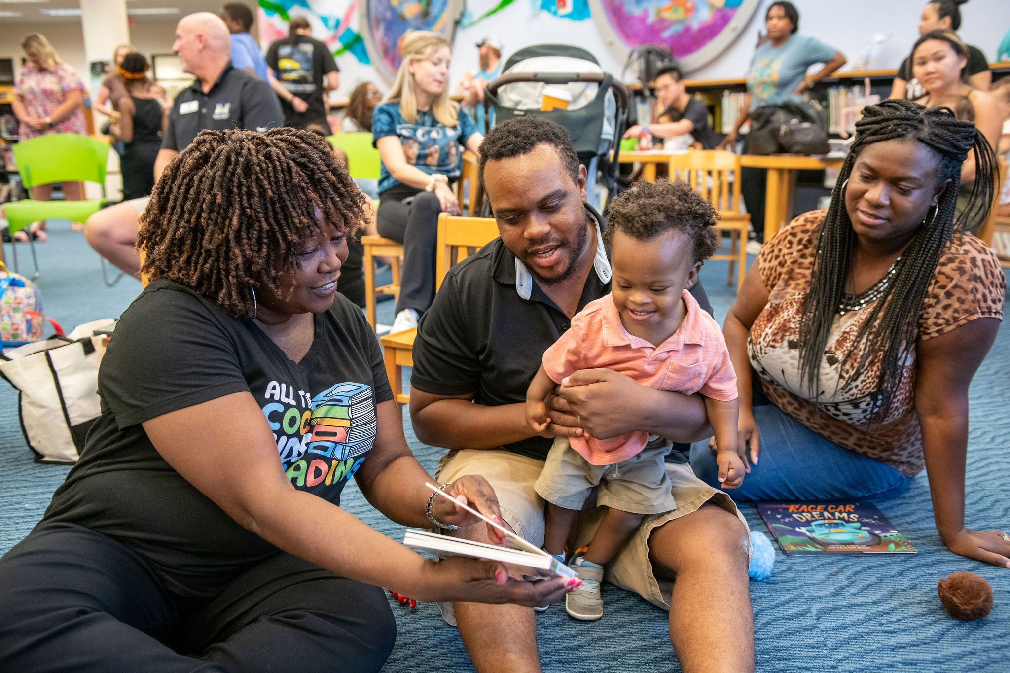 A group of adults and a toddler sit on a carpeted floor in a library, with one woman reading a book to the child while others watch and interact nearby.