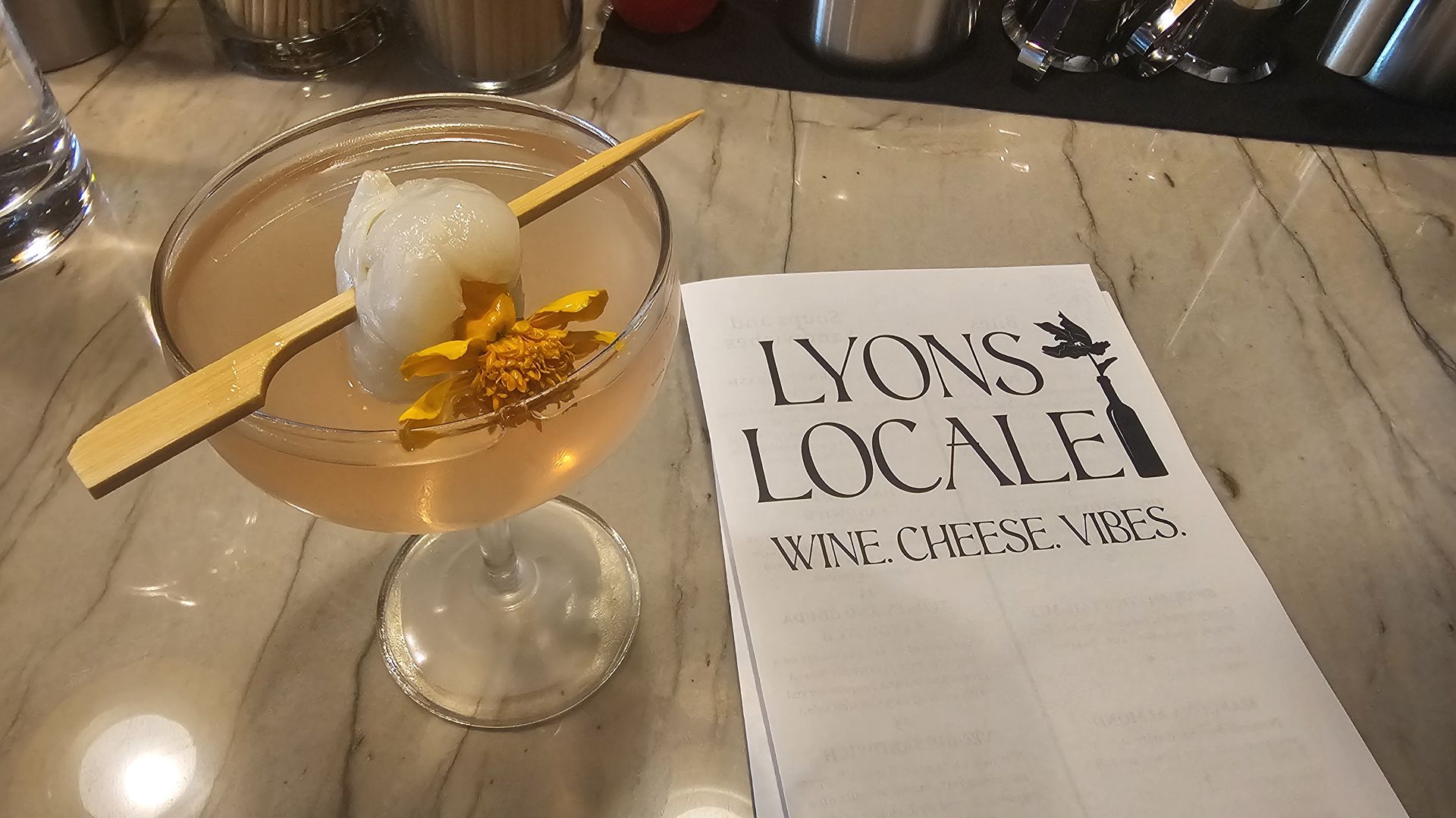 Pink cocktail in a coupe glass garnished with a lychee and yellow flower on a wooden pick, beside a white menu labeled "Lyons Locale, Wine. Cheese. Vibes." on a marble countertop.