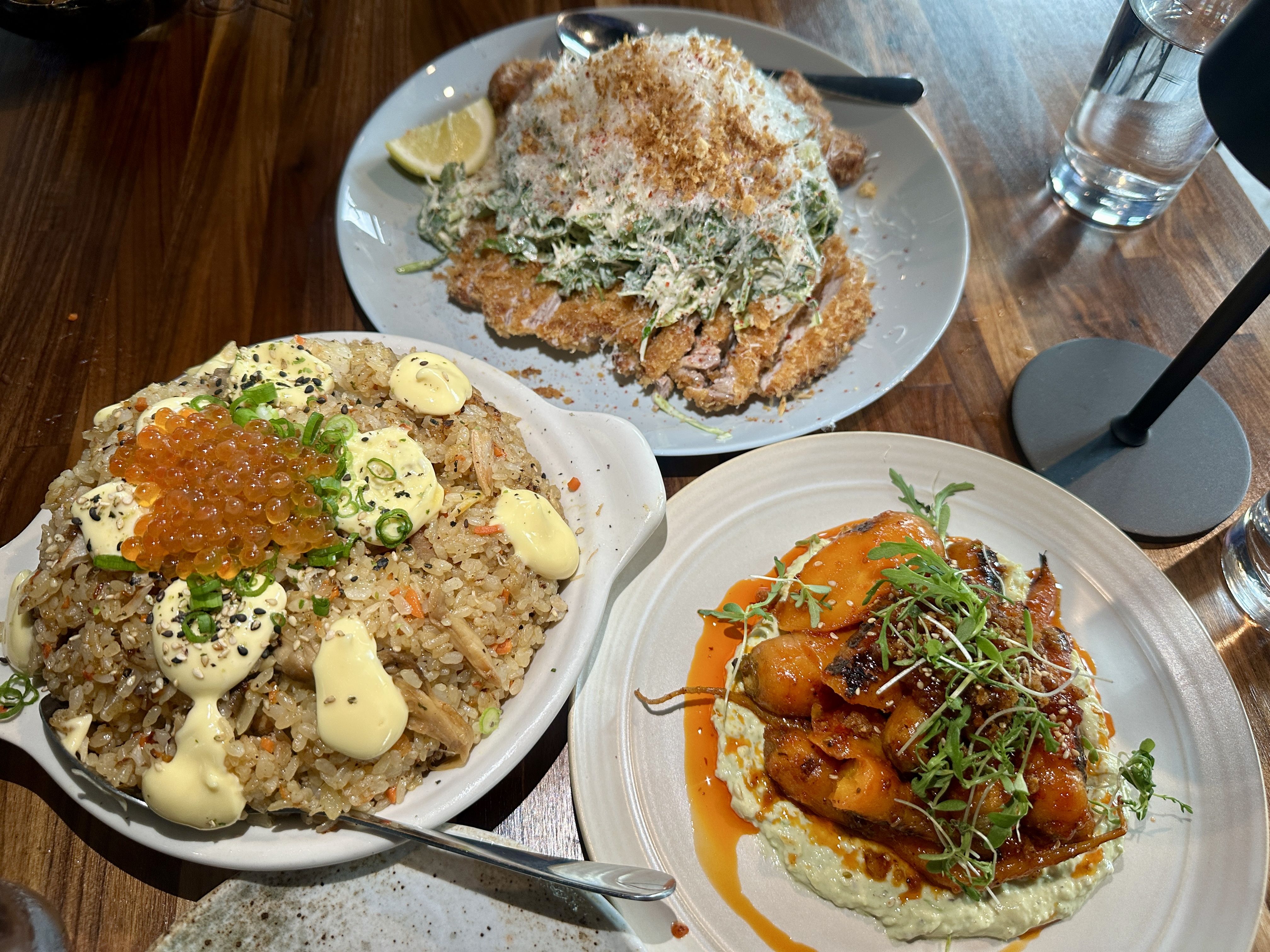 Top-down view of a table with three plates: crispy chicken katsu with shredded cabbage and lemon wedge, fried rice with salmon roe, mayo, sesame, and chives, and a meat dish in orange sauce.