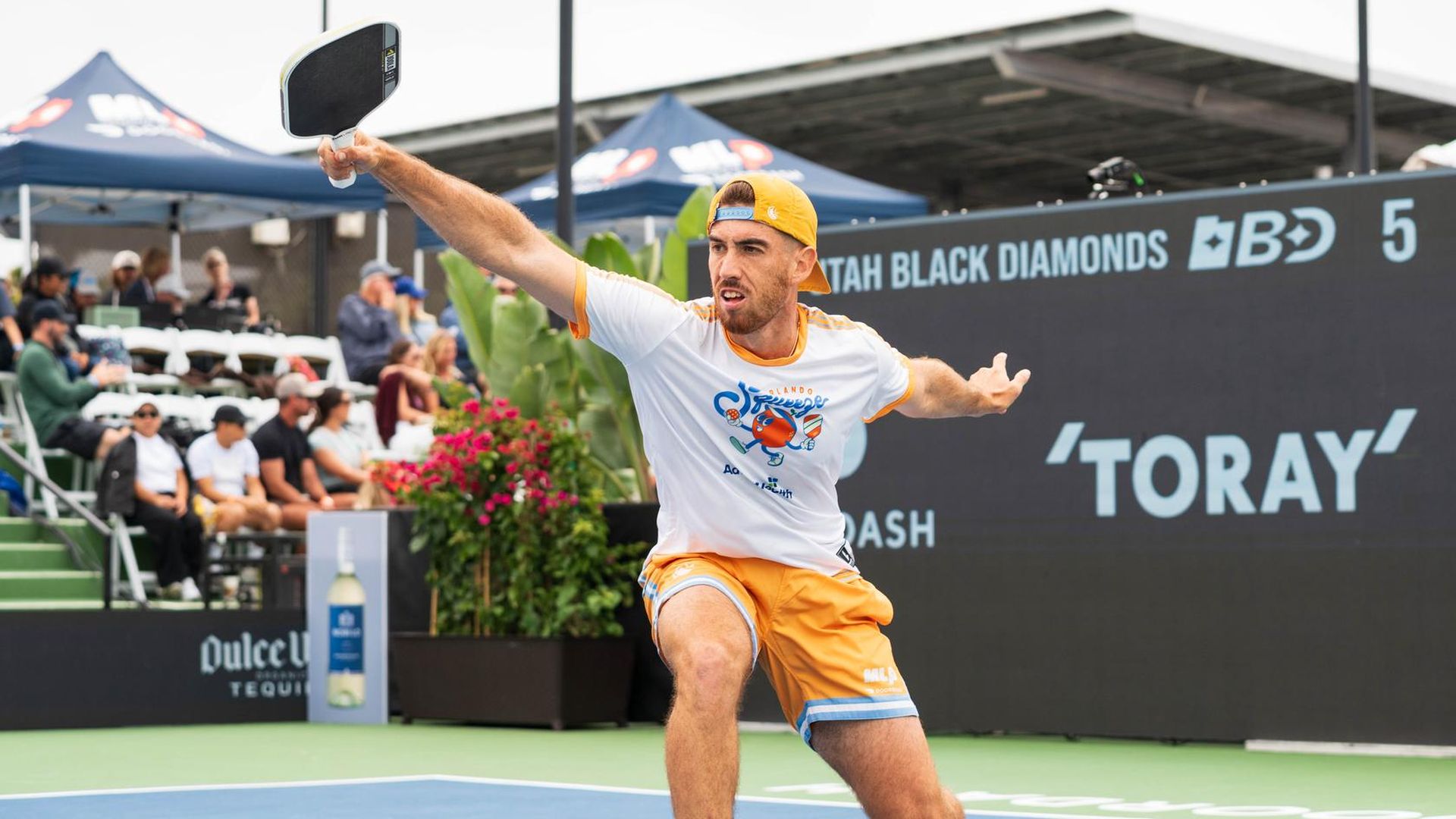 Male pickleball player in a white and orange outfit with a backward orange cap lunges on a blue court, paddle raised. Behind him, spectators in bleachers under tents and a large sponsor board.