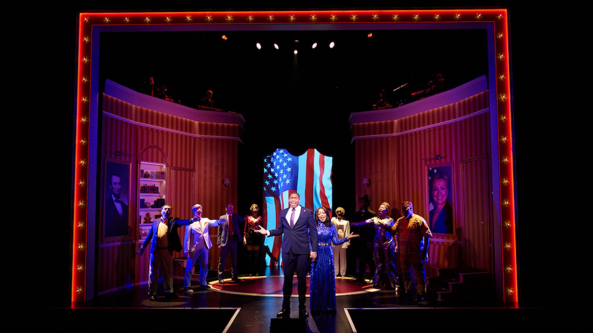 Stage framed by a red bulb-lit border. A cast in formal wear forms a semicircle around a man in a suit and a woman in a blue gown at center, with a waving American flag backdrop.