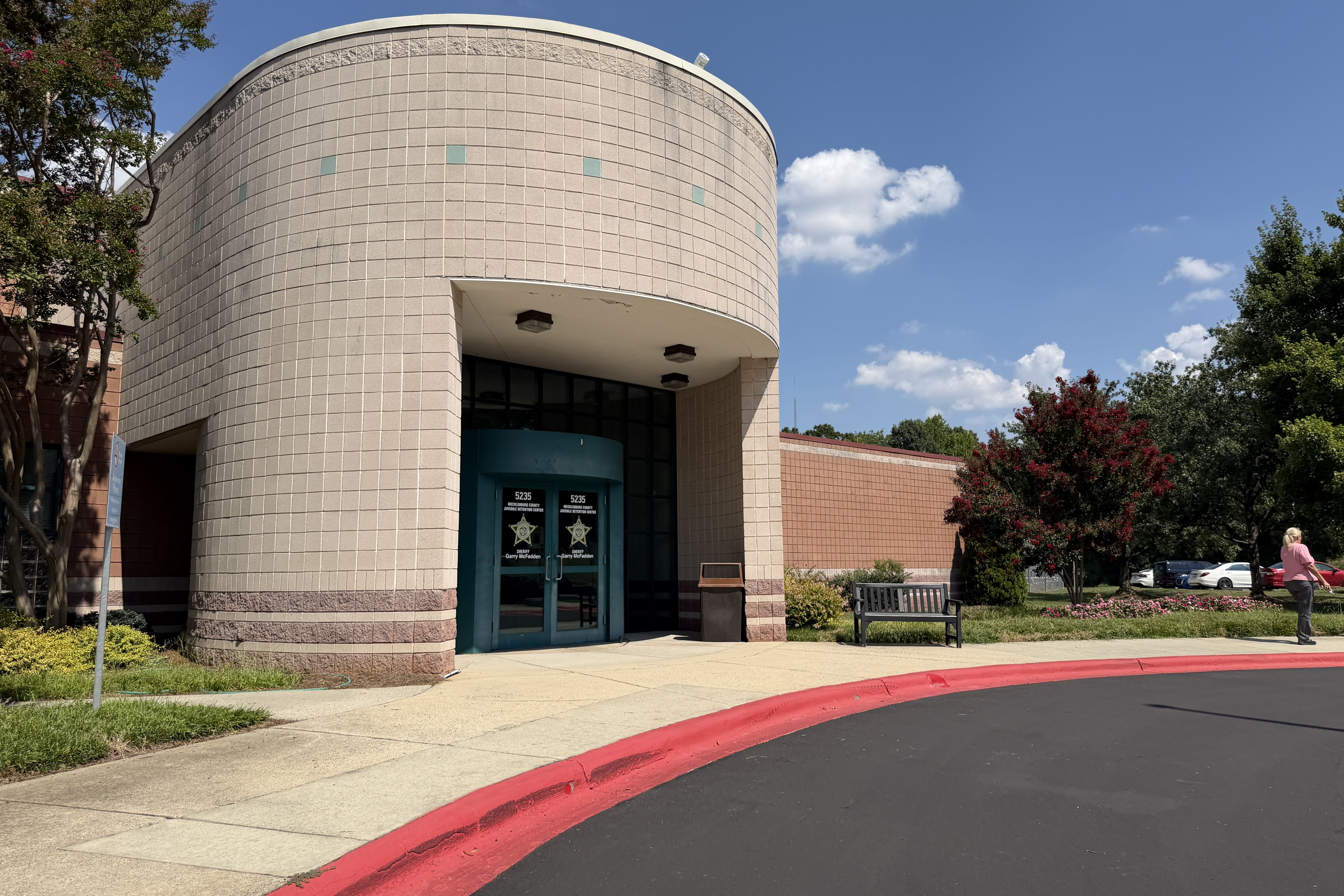 Curved beige tile building with teal doors under a white canopy, set against a blue sky. Trees line the sides; a person walks along a red curb with parked cars nearby.