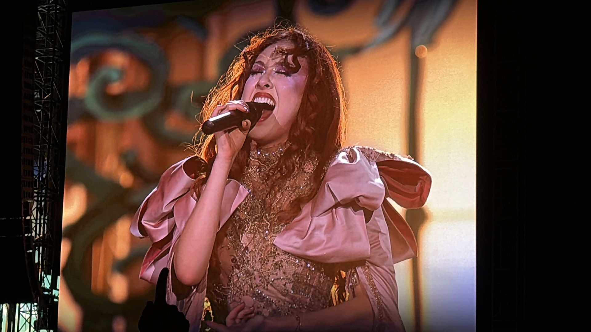 Female singer with curly red hair passionately singing into a microphone, wearing an ornate gold and pink dress with large pink bows, against a warm, blurred background.