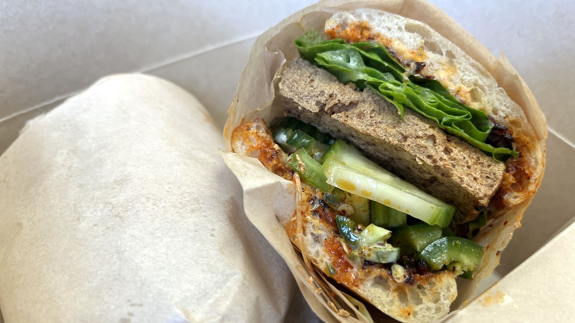 Close-up of a half wrap filled with green lettuce and spinach, cucumber sticks, and brown crusty bread, smothered in orange sauce, wrapped in parchment.
