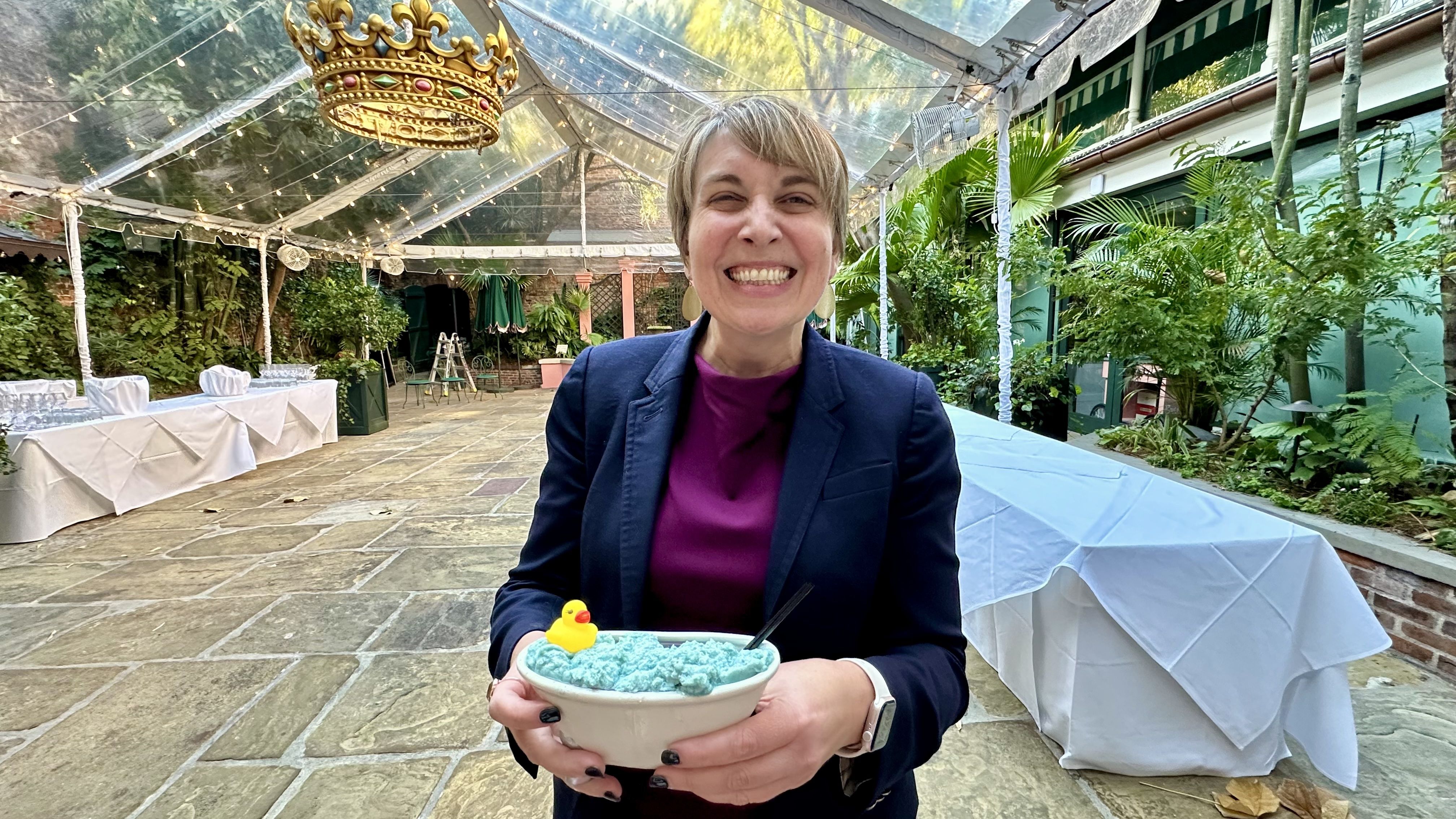 Smiling woman in a navy blazer and purple top holds a cocktail with a yellow rubber duck topper, sitting in a covered outdoor area with greenery and tables covered in white cloth.