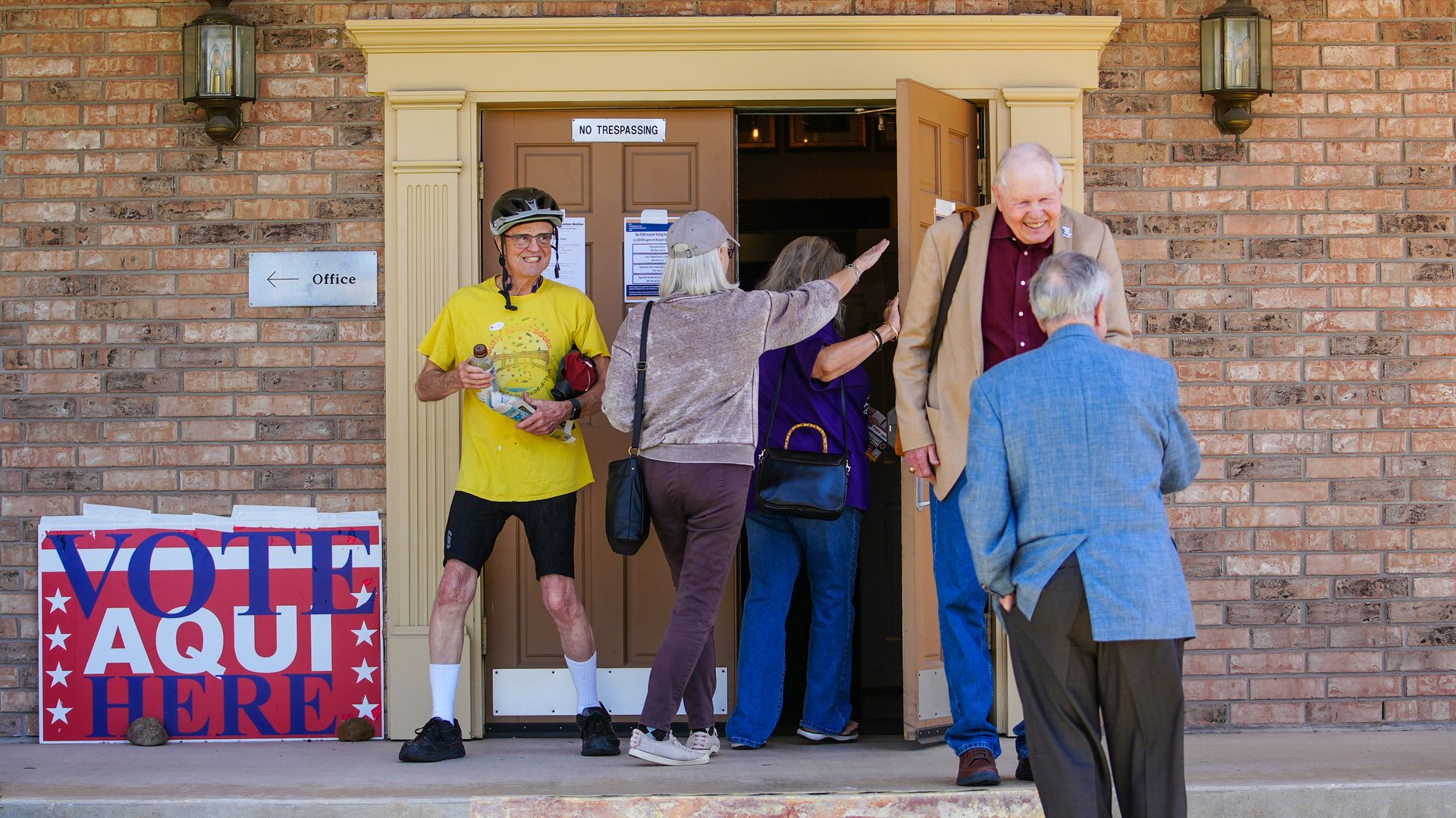 Brick building entrance with beige doorway and two lanterns. People stand at an open door labeled "No Trespassing"; a man in a yellow shirt and helmet holds a bottle. A "VOTE HERE" sign sits nearby.