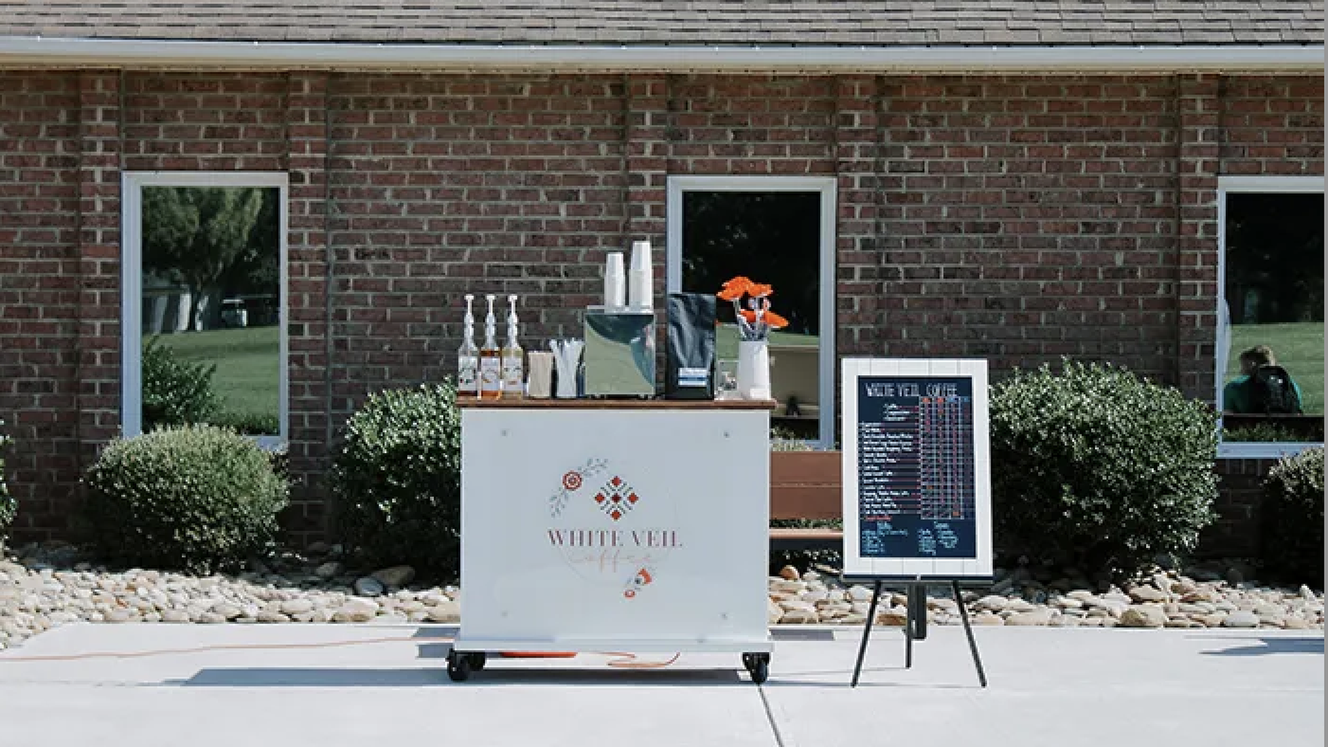 Outdoor scene with a white mobile beverage cart on wheels in front of a brick building, bottles, cups, an ice bucket and orange flowers, beside a chalkboard menu and shrubbery.
