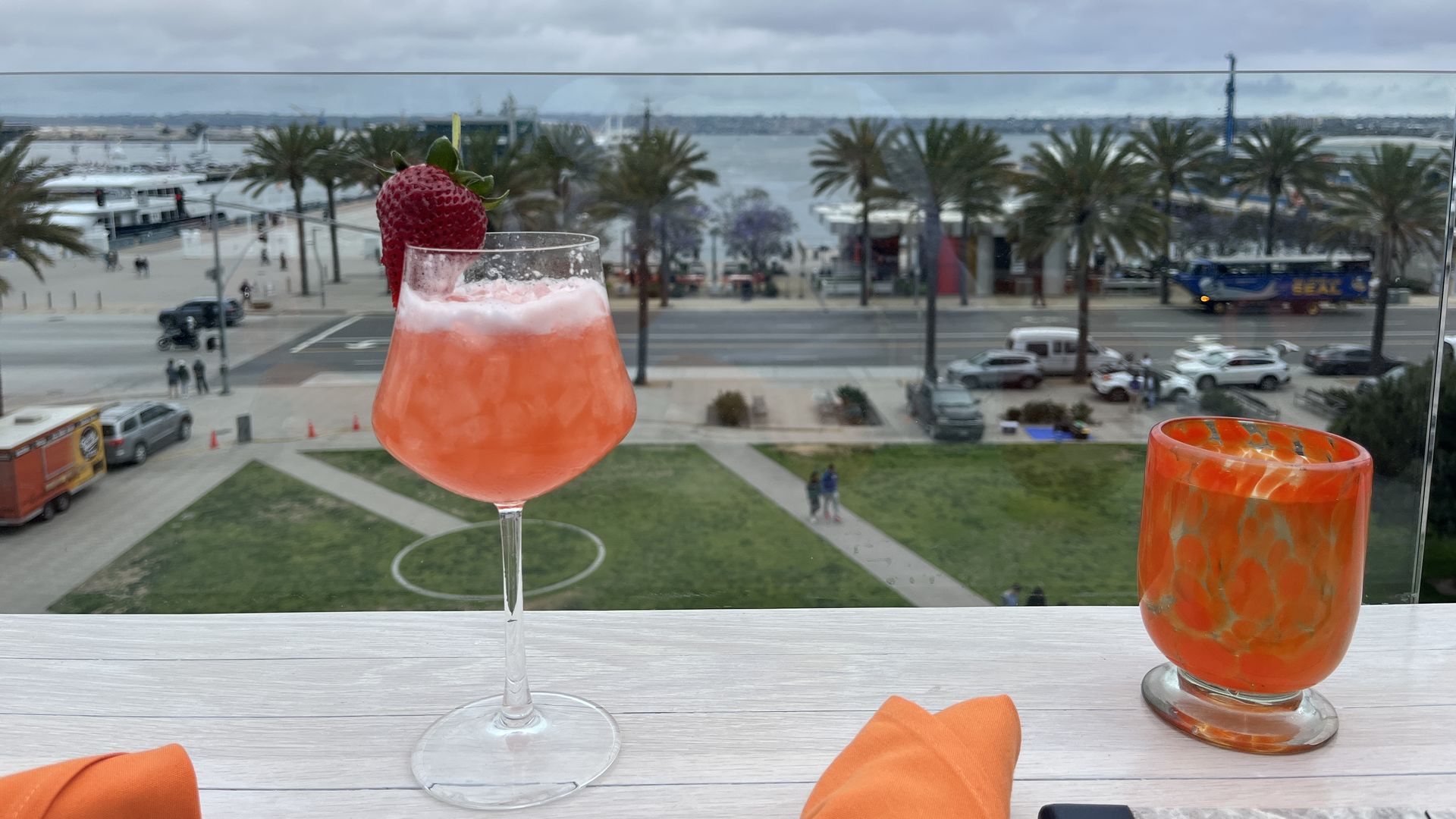 An orange-colored cocktail sits on a ledge at a rooftop bar overlooking a grassy park, palm trees and the San Diego Bay.