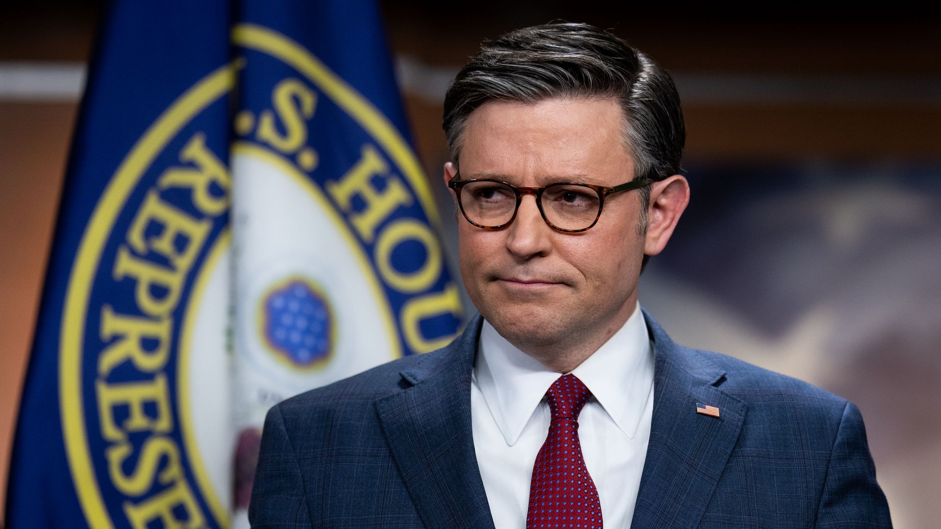 House Speaker Mike Johnson, wearing a blue suit, white shirt and red tie, standing in the Capitol's studio A in front of a U.S. House of Representatives flag.