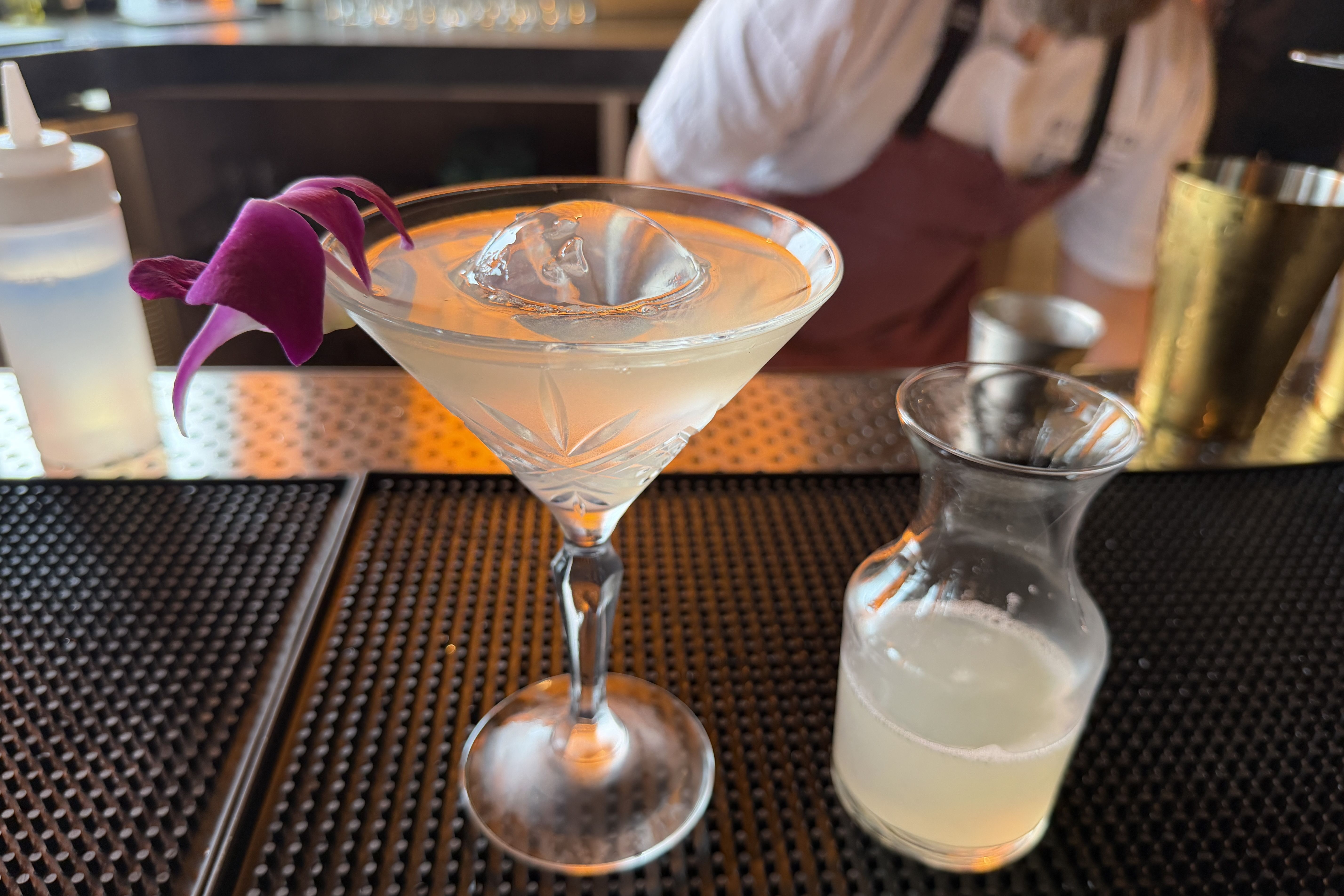 Martini glass with a large ice cube and a purple orchid garnish sits on a bar counter; a blurred bartender in a white shirt and maroon apron is in the background.