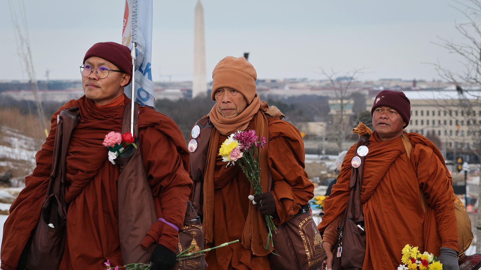 Three Buddhist monks wearing robes and knit hats carry flowers on a walk near Washington, D.C. The Washington Monument can be seen in the background. 