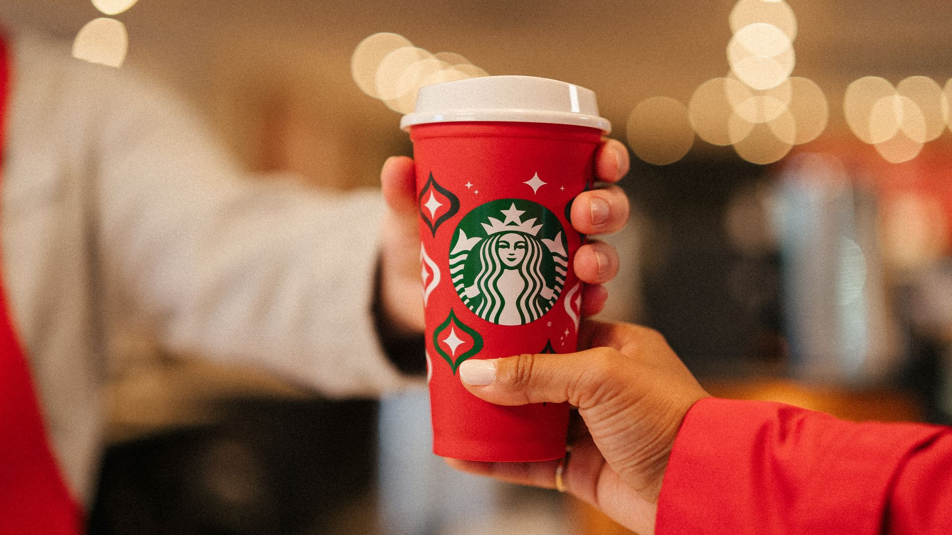 Two hands (one wearing beige shirt and other red shirt) holding a red Starbucks cup