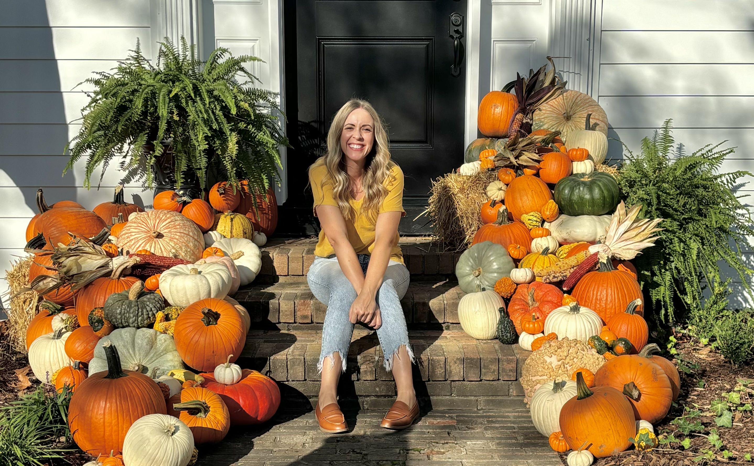 Smiling woman in a yellow shirt and jeans sits on brick steps surrounded by various pumpkins and gourds in orange, white, and green, with green ferns on a sunny porch entrance.
