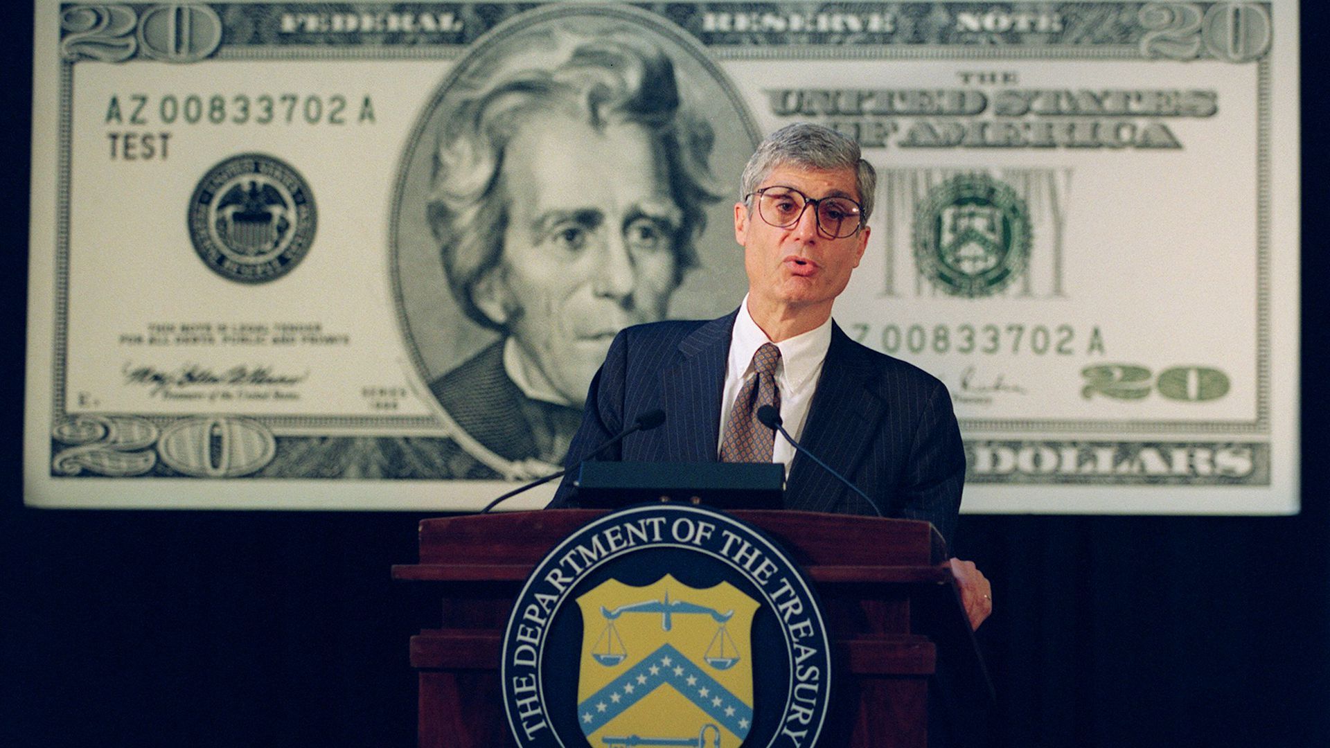Photo of a bespectacled person wearing a suit and speaking from a podium
