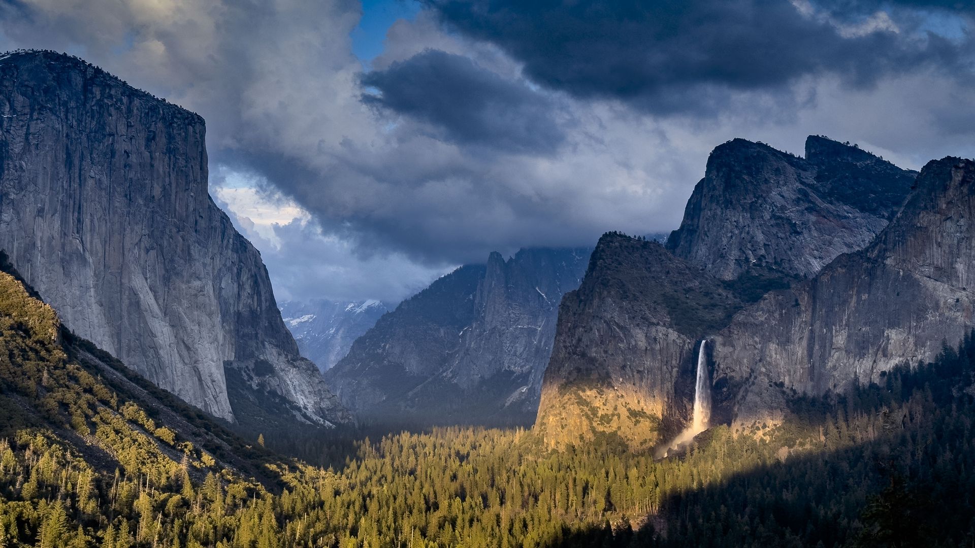 The sun shines on Yosemite Valley trees as dark clouds linger over mountains and a waterfall. 