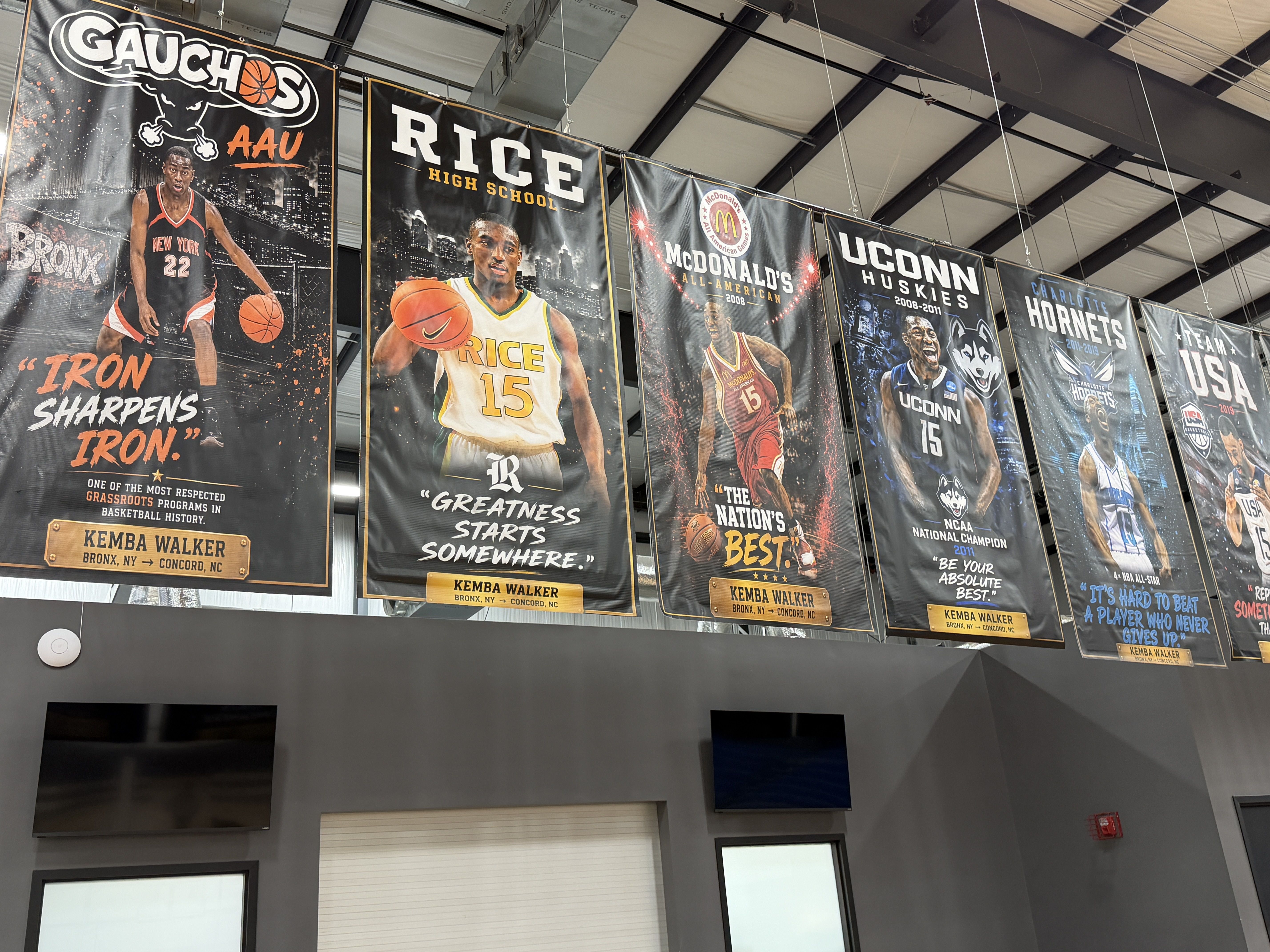 Row of large vertical basketball banners hanging from a high gym ceiling, featuring players and team names such as Gauchos, Rice High School, McDonald's All-American, and UConn Huskies.