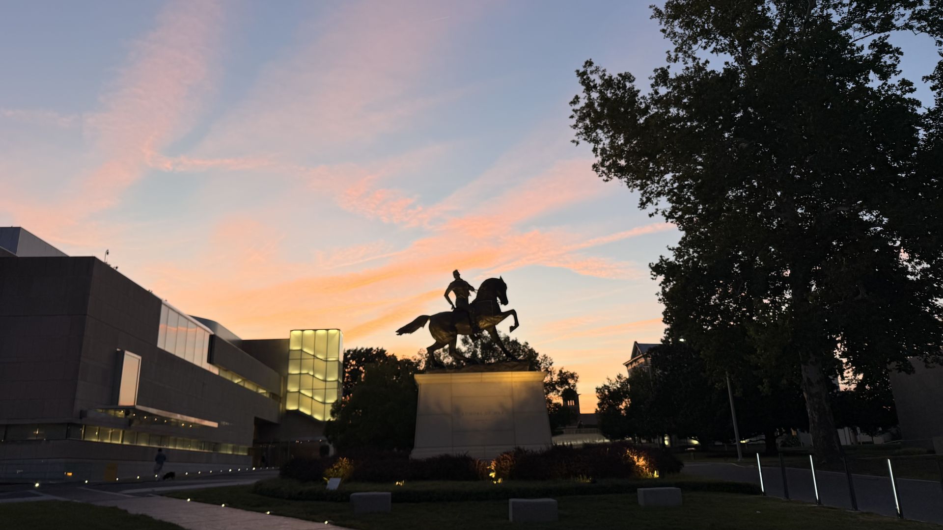 Silhouette of an equestrian statue on a pedestal in a city plaza at sunset, with pink-orange clouds, modern buildings to the left and a large tree to the right.