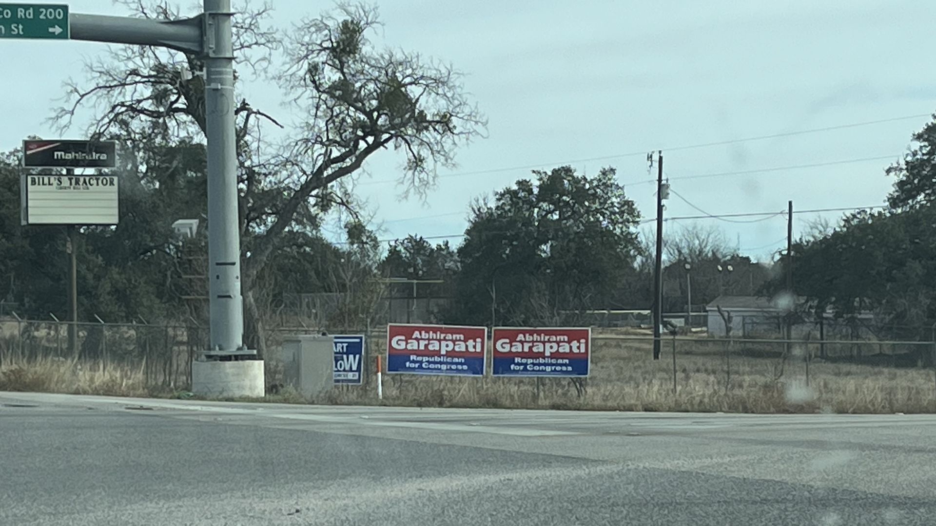 Two red and blue political signs for Abhiram Garapati, Republican for Congress, on grassy roadside near a traffic light and a sign for Bill's Tractor under a cloudy sky.