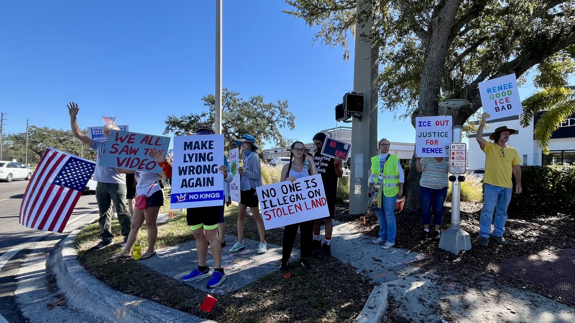 Group of protesters standing on a street corner holding signs with messages such as "WE ALL SAW THE VIDEO," "MAKE LYING WRONG AGAIN," "NO ONE IS ILLEGAL ON STOLEN LAND," and "RENEE GOOD ICE BAD" under a clear blue sky.