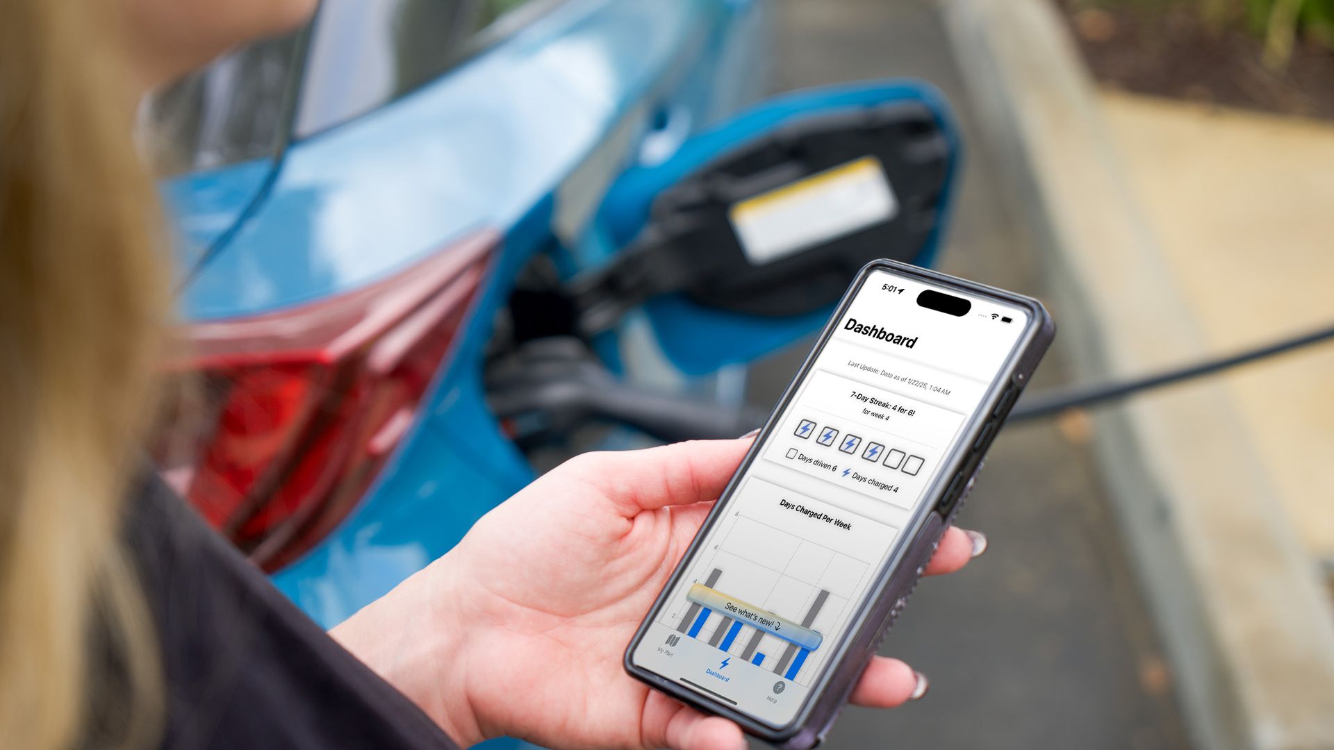 Person holding a smartphone displaying a dashboard app while charging a blue electric vehicle plugged into a charging station outdoors.