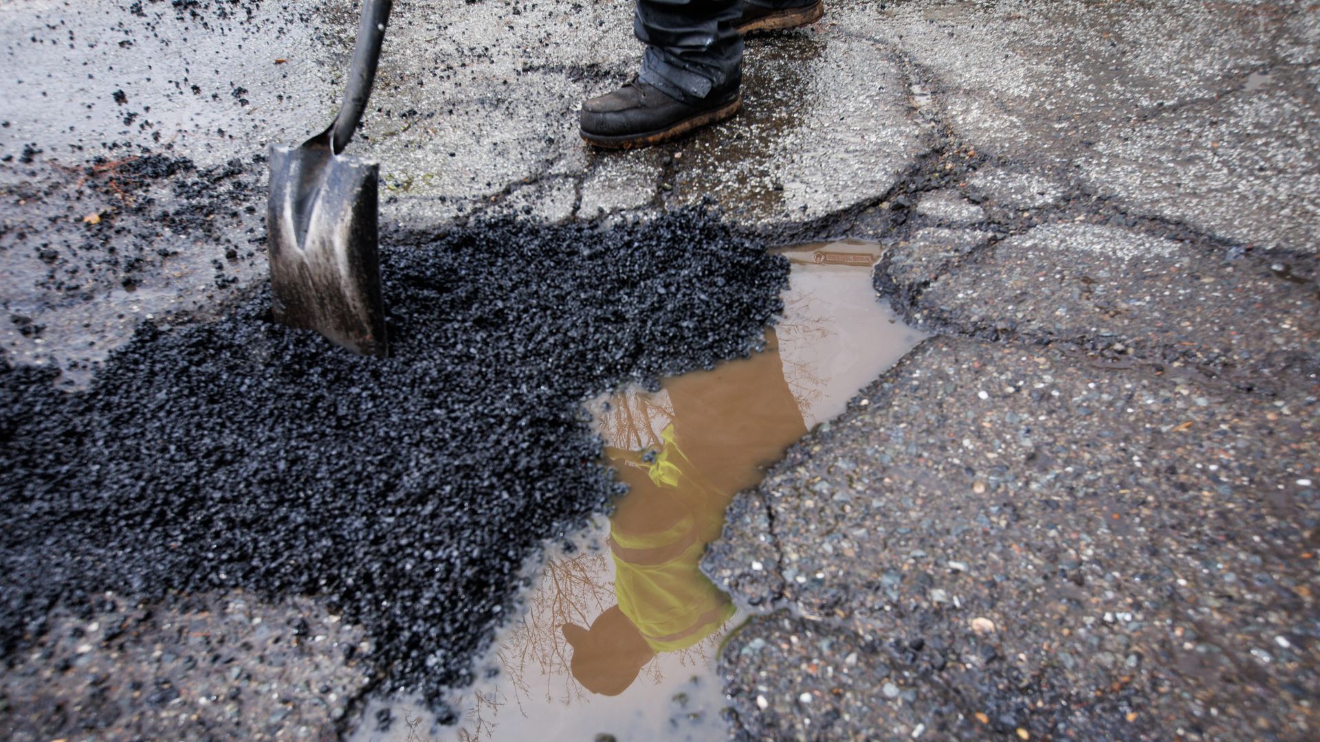 A shovel and a brown shoe belonging to a maintenance worker are shown above a pot hole filled with brown water. The maintenance worker's reflection is visible in the puddle.
