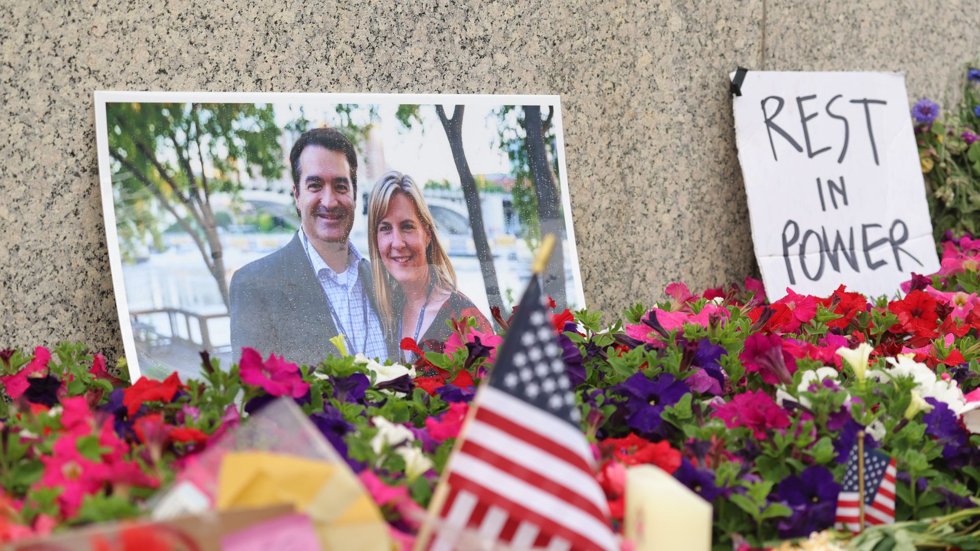 Memorial of colorful flowers, American flags, and a photo of a smiling man and woman (Mark and Melissa Hortman) near a granite wall with a sign reading "REST IN POWER."