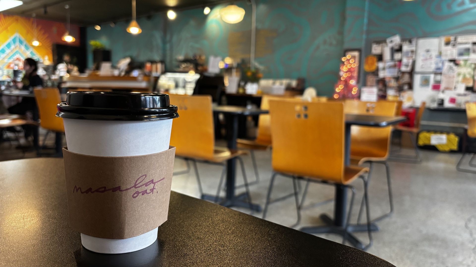 White takeaway coffee cup with black lid and brown sleeve on a table in a cafe with orange chairs, teal wall, and bulletin board covered in notes and pictures.