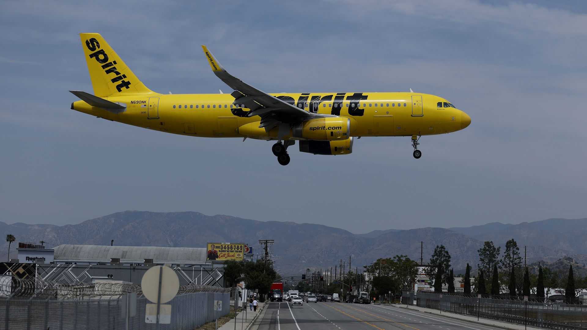 Bright yellow Spirit Airlines jet flies low over a city street near an airport, with chain-link fences, buildings, cars below, and distant mountains under a blue sky.