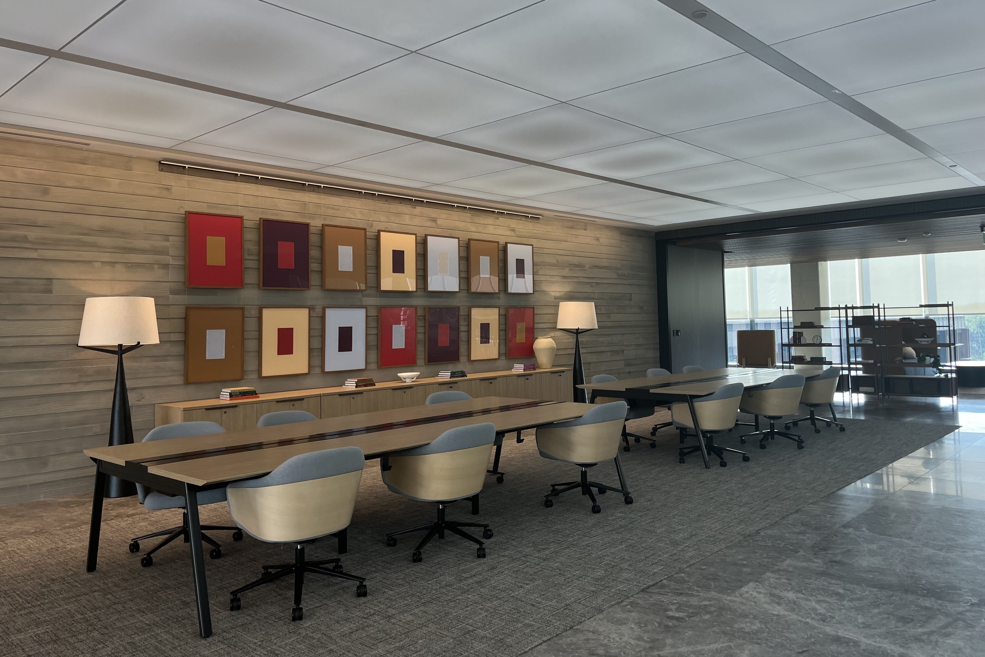 Modern conference room with long wooden tables, gray and beige chairs on wheels, wall art consisting of colorful framed rectangles, two tall lamps, and a shelf unit near large windows.