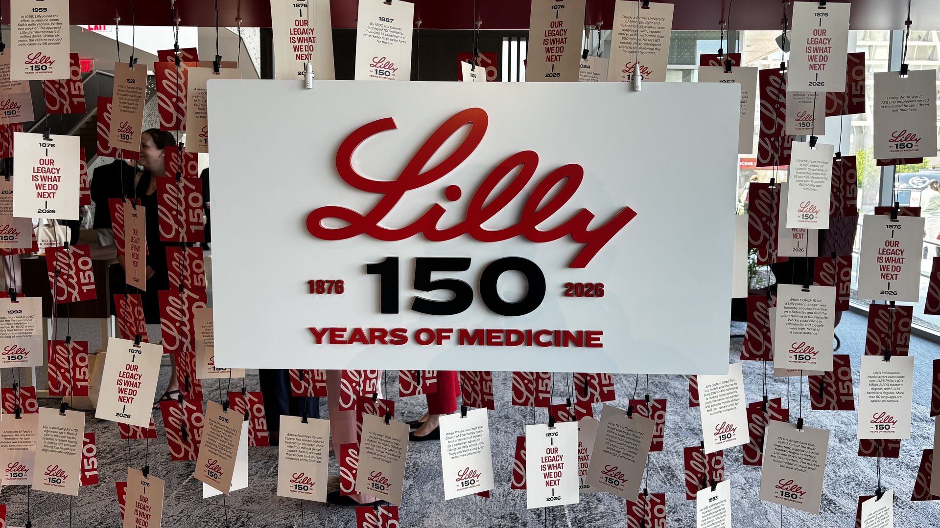 Large white display featuring the red Lilly script logo and the text "150 Years of Medicine" with the years 1876–2026; surrounded by hanging red-and-white Lilly 150 cards, and a person visible in the background.