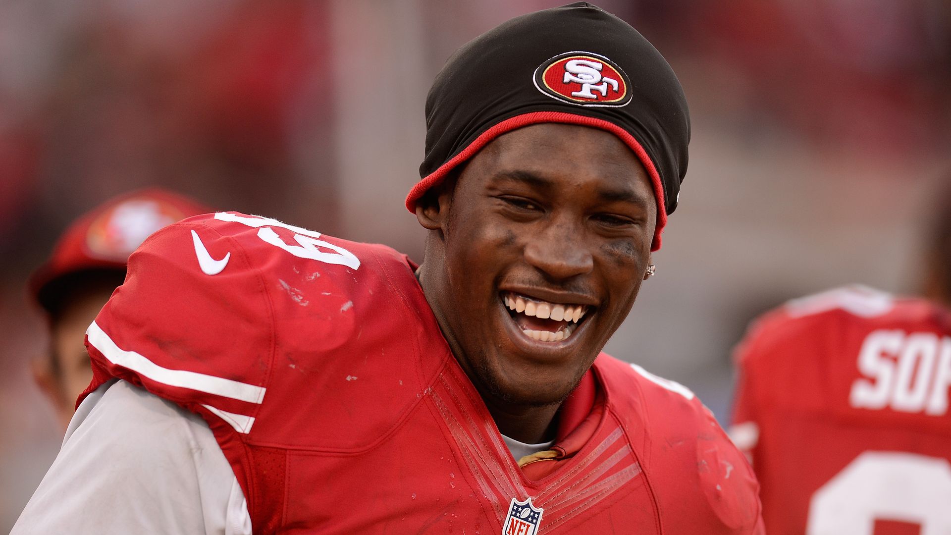 Smiling football player wearing a red 49ers jersey and a black beanie with the team logo; dirt smudges on the uniform, blurred stadium crowd in the background.