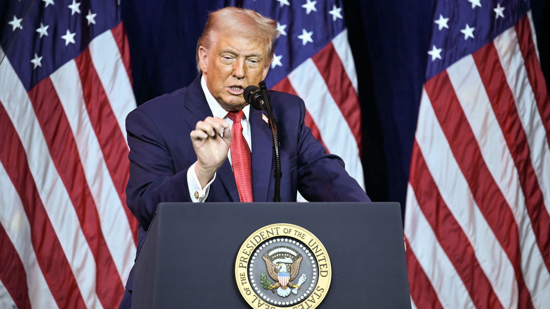 President Trump, in a dark suit, white shirt, and red tie, speaks at a podium with the Seal of the President of the United States, with American flags in the background.