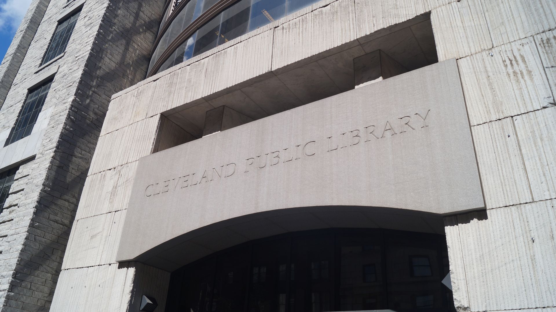 Exterior of the Cleveland Public Library, Louis Stokes Wing. Gray stone walls, tall windows, and a curved glass facade above a wide concrete entry, under a blue sky.