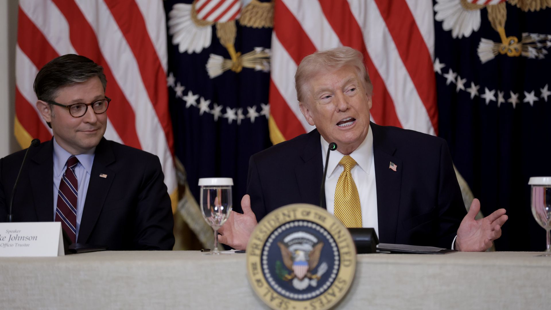 Two men in suits at a panel, American flags behind. The man on the right in a dark suit and yellow tie speaks into a microphone; a presidential seal sits on the table, with water nearby.