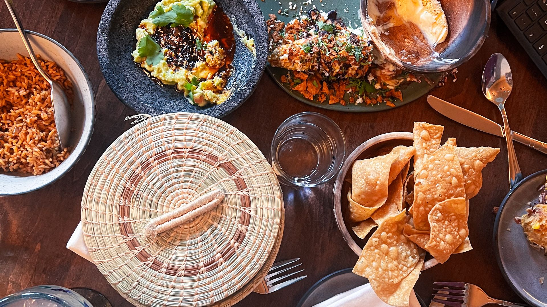 Top-down view of a wooden table loaded with Mexican-inspired dishes: bowls of rice, guacamole, salsa, shredded meat, tortillas and chips, lime, cilantro, hot sauce, and a can of Coke.