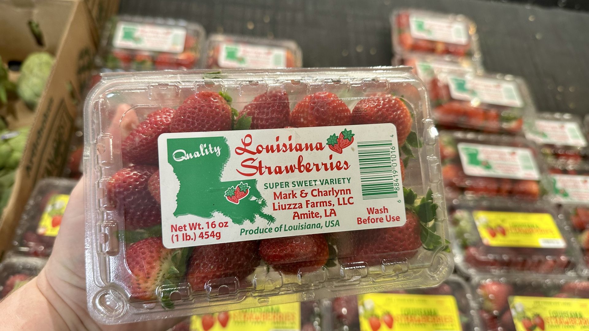 Clear plastic clamshell of fresh red strawberries held by a hand in a store aisle. Label reads "Louisiana Strawberries" with a green map of Louisiana, barcode, and "Wash Before Use" on the label.