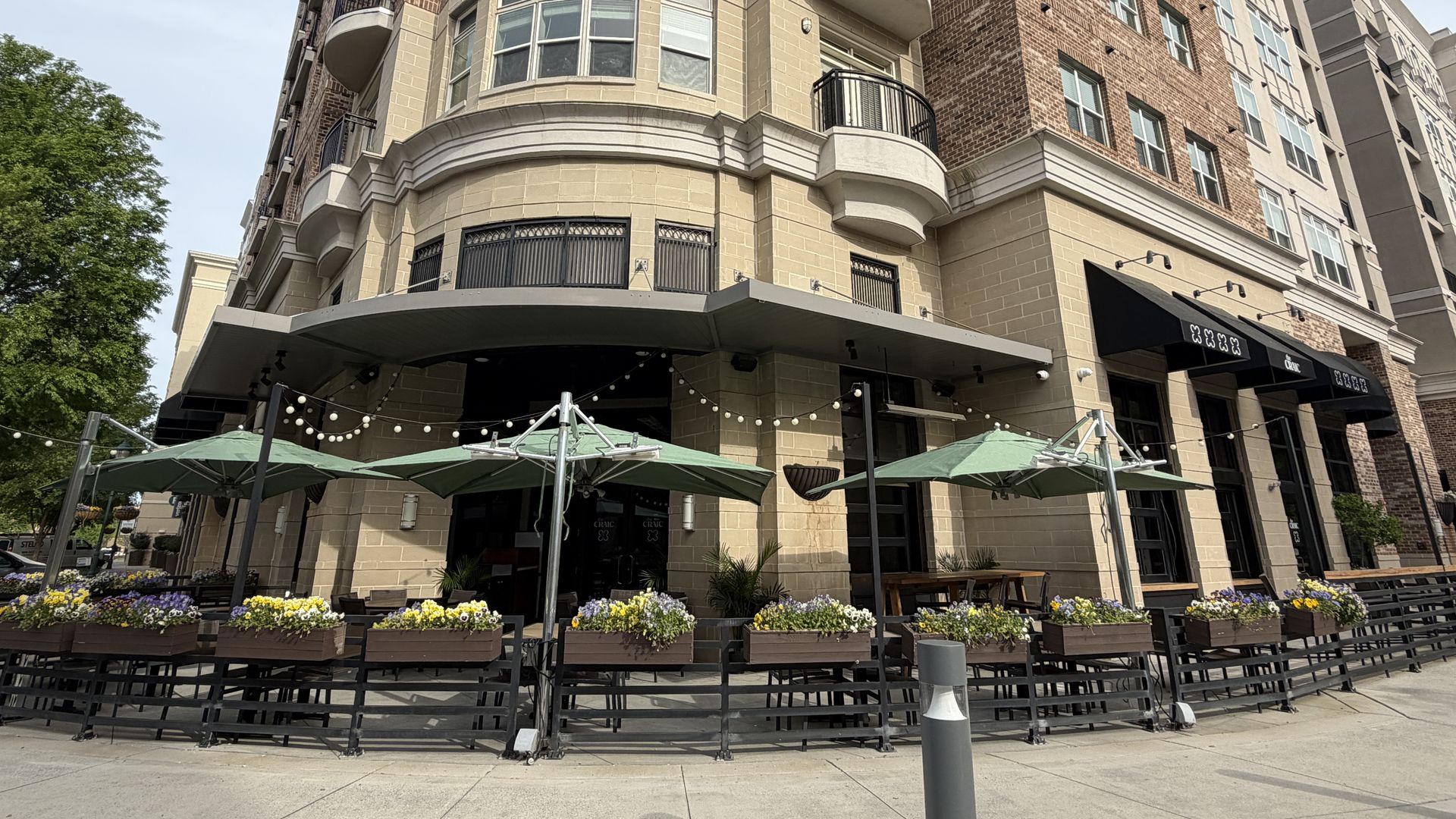 Multi-story brick building with curved balconies and beige stone base. Ground-floor shops with black awnings, outdoor seating under green umbrellas, string lights, and flower planters along sidewalk.