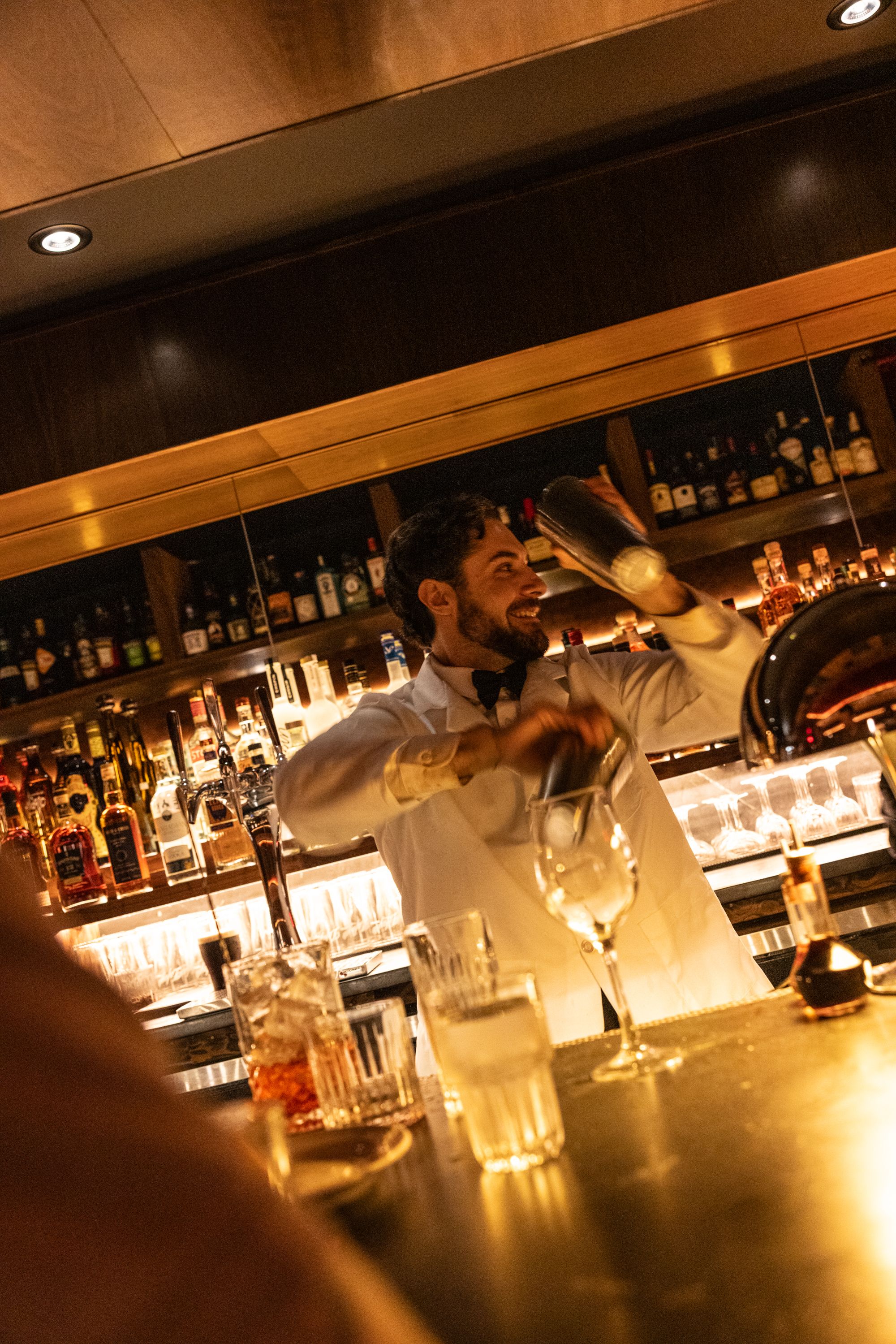 A smiling bartender in a white shirt and black bow tie shakes a cocktail shaker behind a warmly lit bar. Shelves of liquor bottles and glassware line the back, glowing amber under soft lighting.
