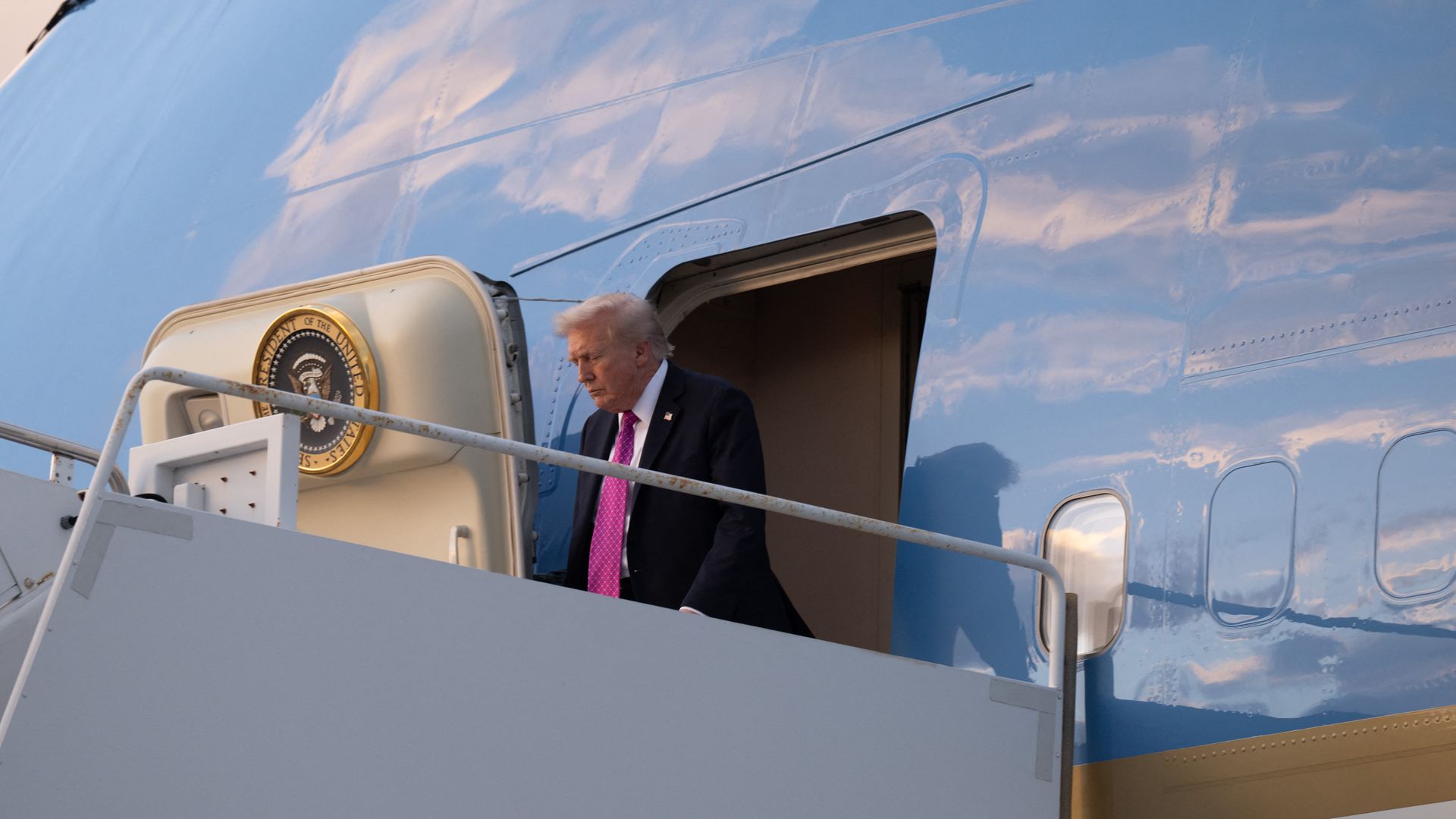 President Trump, wearing a navy jacket with a US flag pin at the top of his left lapel, white shirt and pink tie with white checks, disembarks from the blue Air Force One upon arrival at Palm Beach International Airport in West Palm Beach, Florida.
