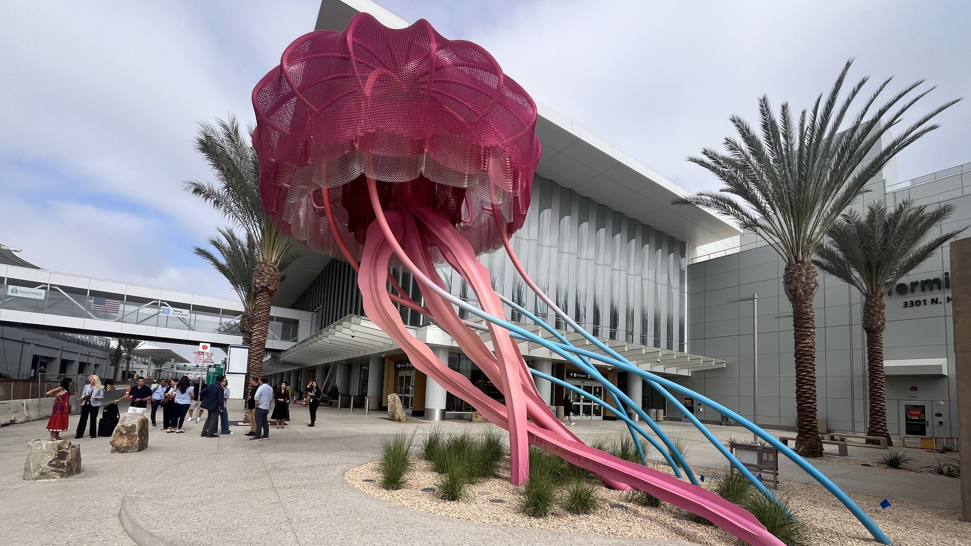 Large pink and blue jellyfish sculpture with long tentacles displayed outside the new Terminal 1 buildings at the San Diego airport with palm trees and people standing nearby.